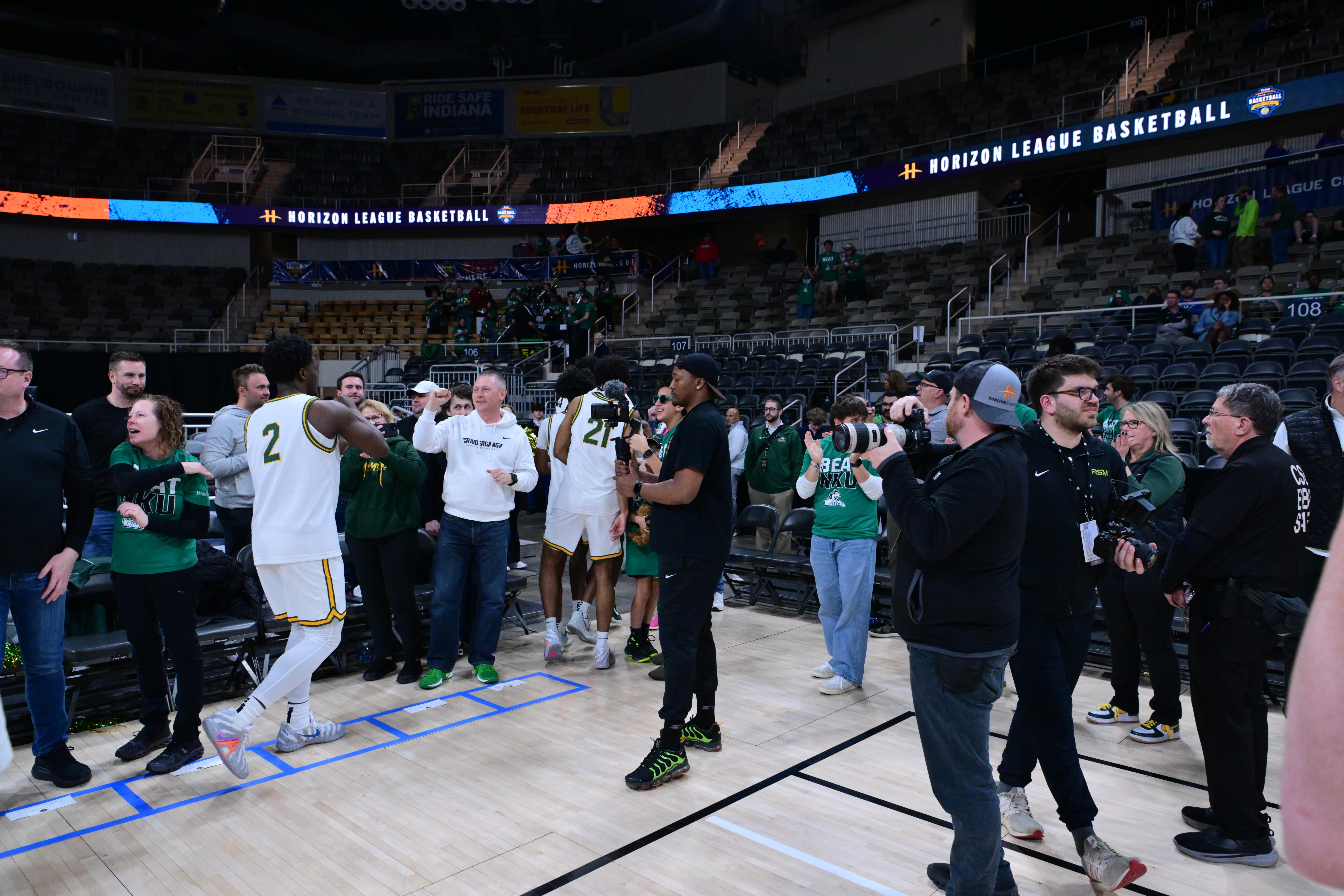 Group of basketball players and staff on court, some clapping and talking, arena seats in background, Horizon League Basketball sign visible.