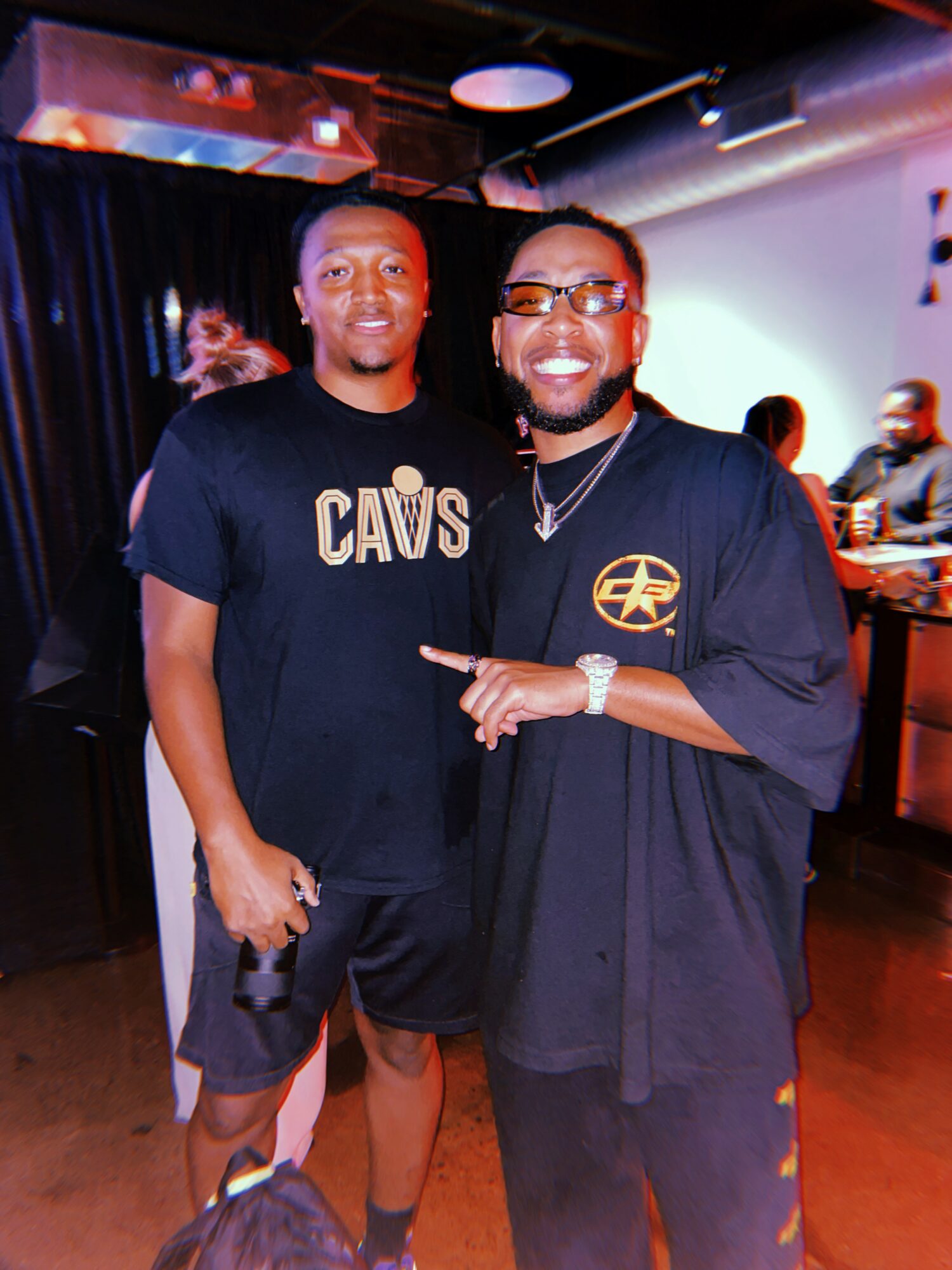 Two men smiling and posing together in a dimly lit indoor setting, one wearing a CAWS t-shirt and the other in a black shirt with a logo, pointing at each other.