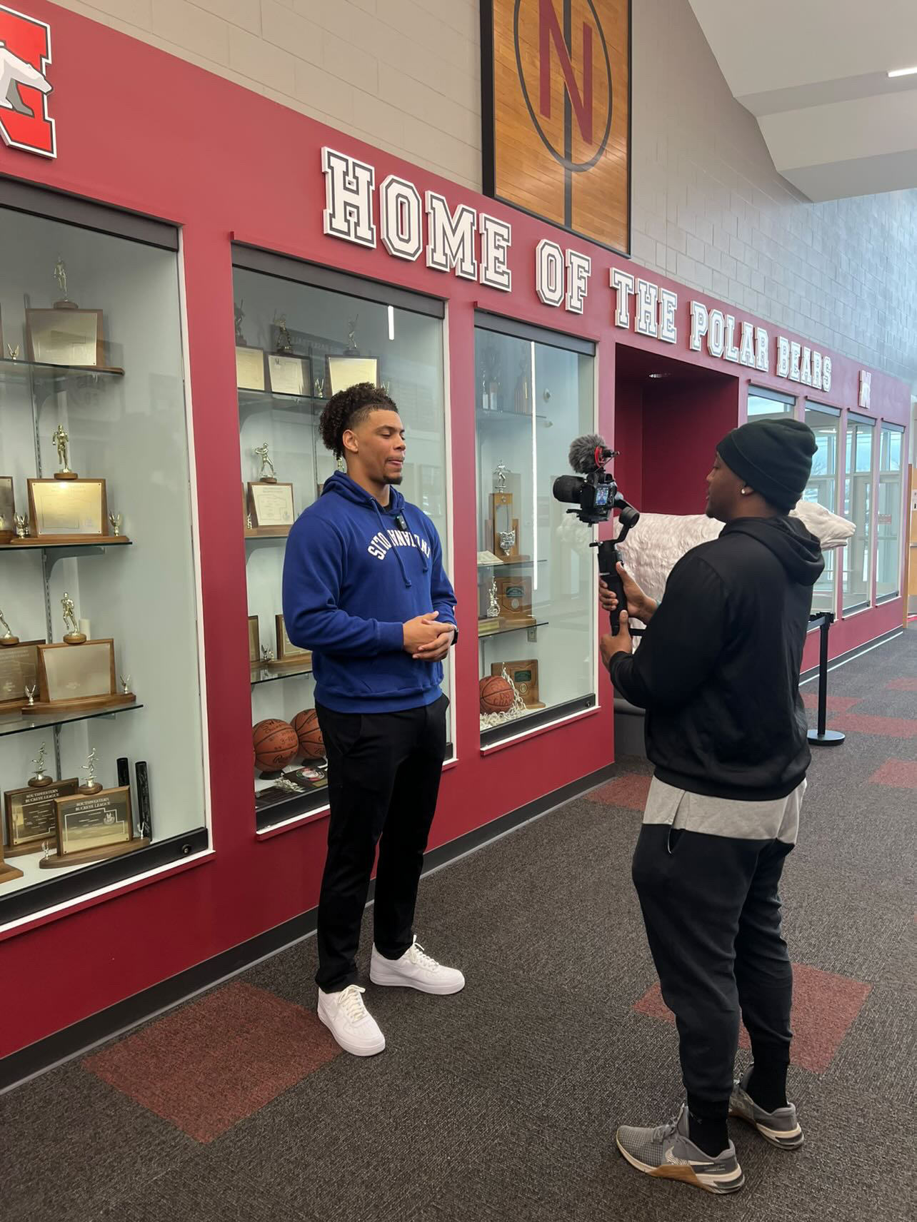 Two people standing in front of display cases with sports memorabilia, one being interviewed or filmed, in a hallway.