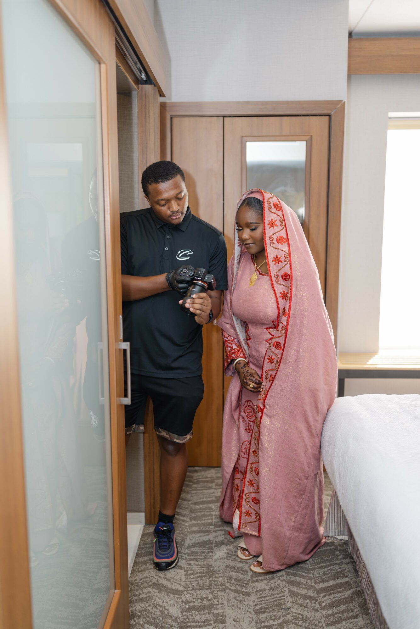 A man and woman stand together in a doorway, looking at a phone, with a bed visible on the right side.