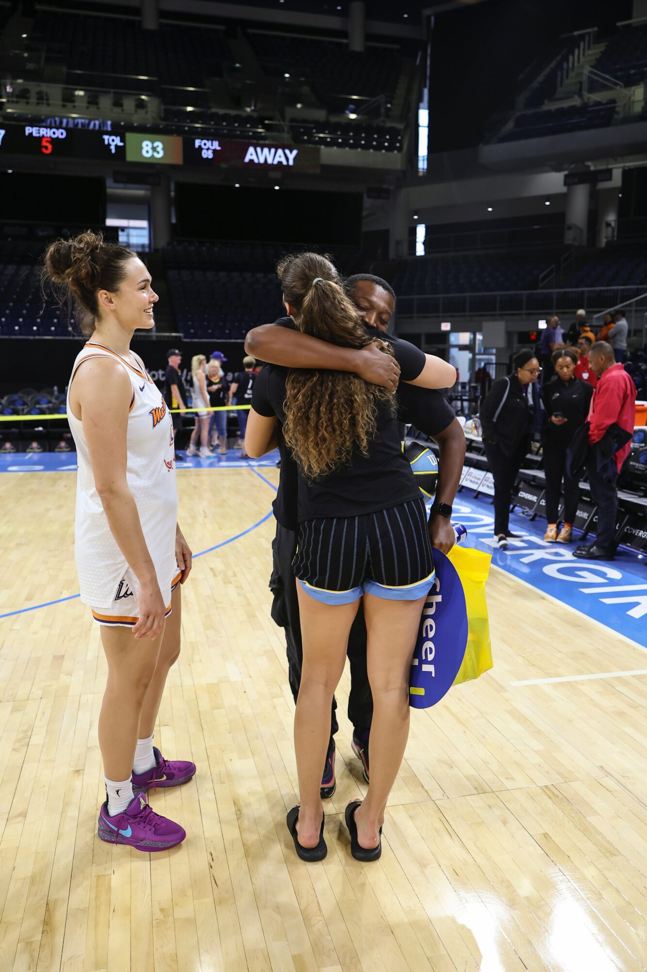 Two female basketball players hugging on the court, one in a black uniform, the other in a white uniform, with a third player nearby.