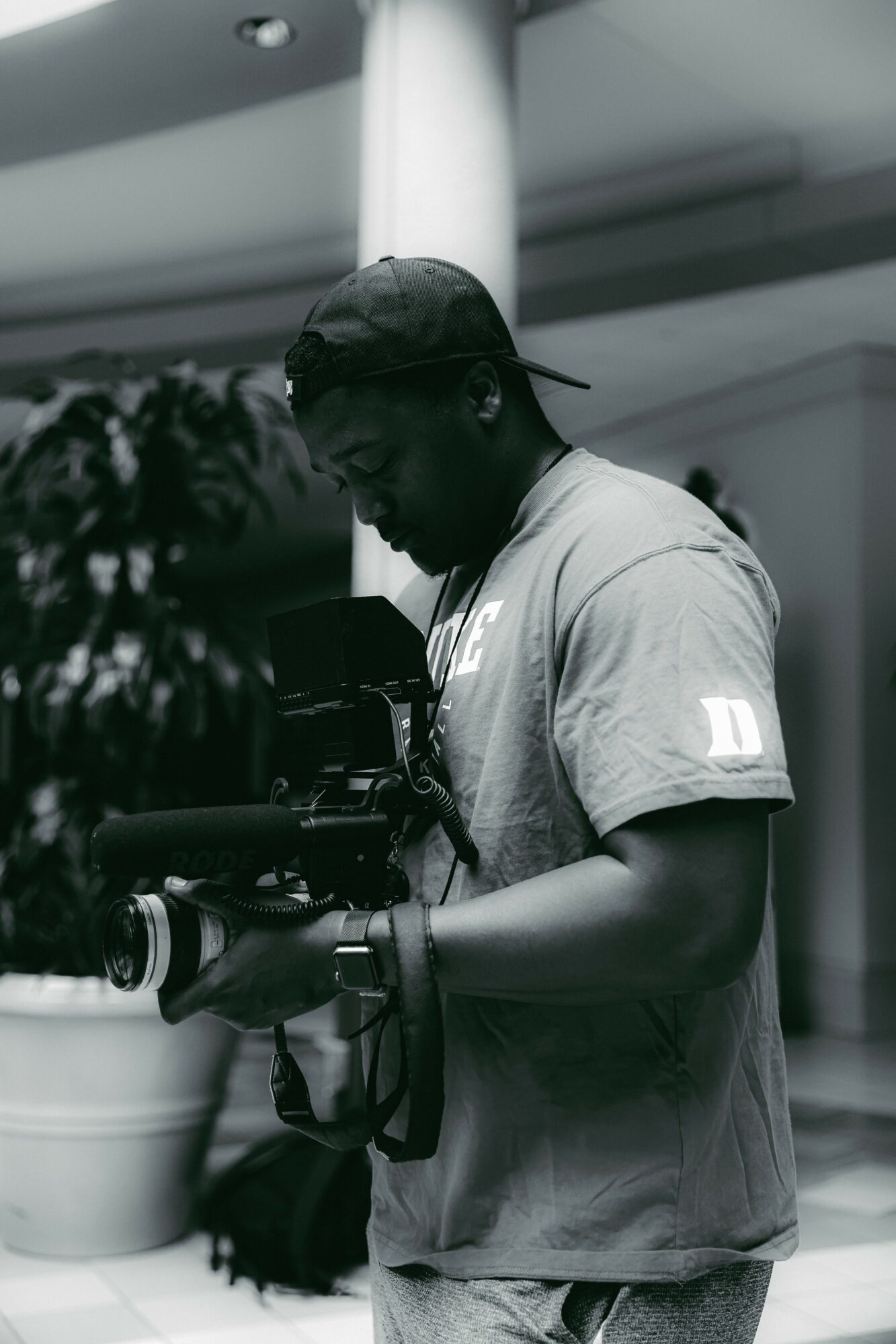Person wearing a cap and glasses holding a camera, standing indoors near a pillar, with a plant nearby.