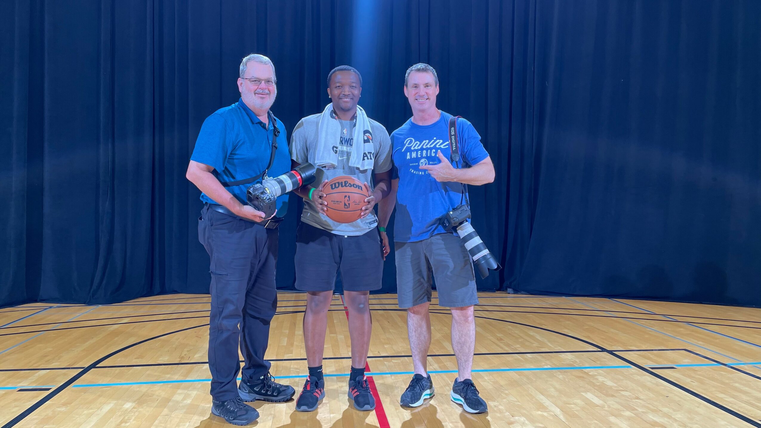 Three people standing on a basketball court with a dark curtain background, holding cameras and a basketball.
