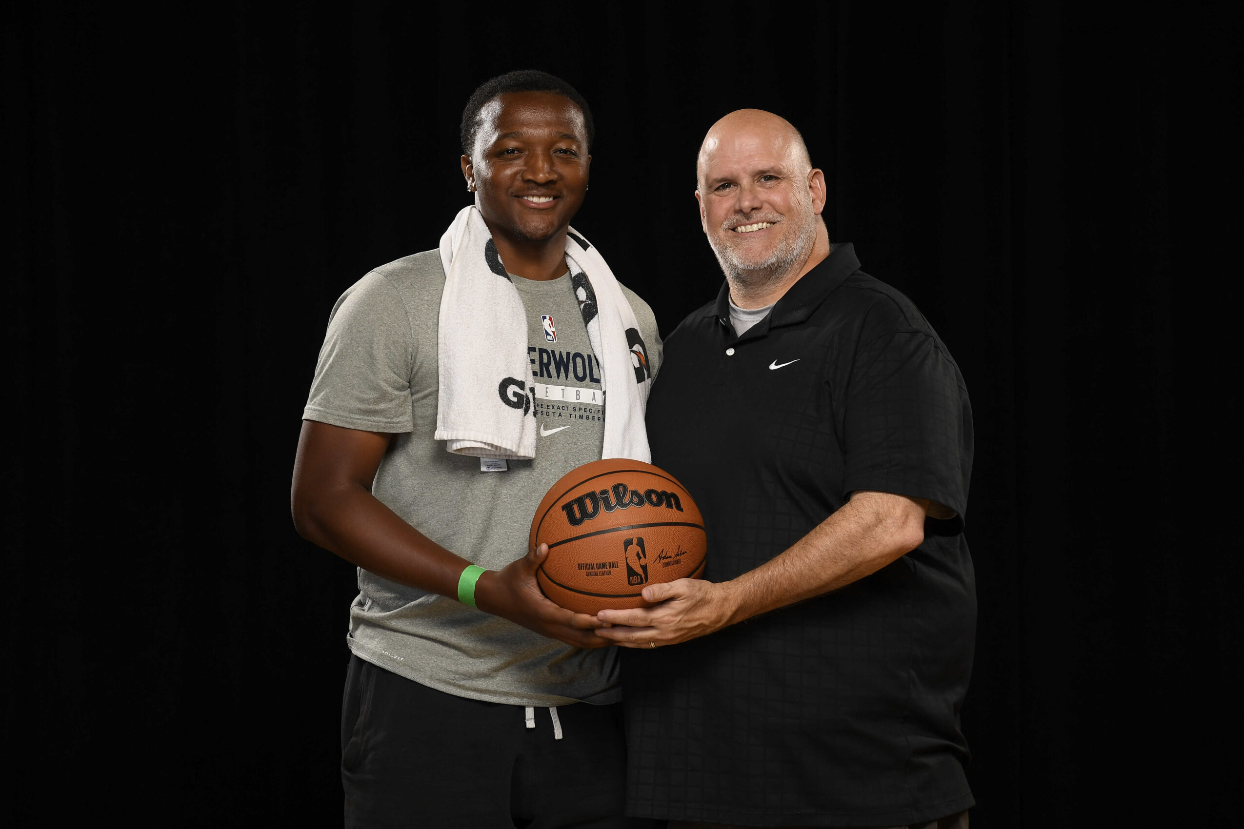 Two men standing together, one holding a basketball, smiling, against a black background.