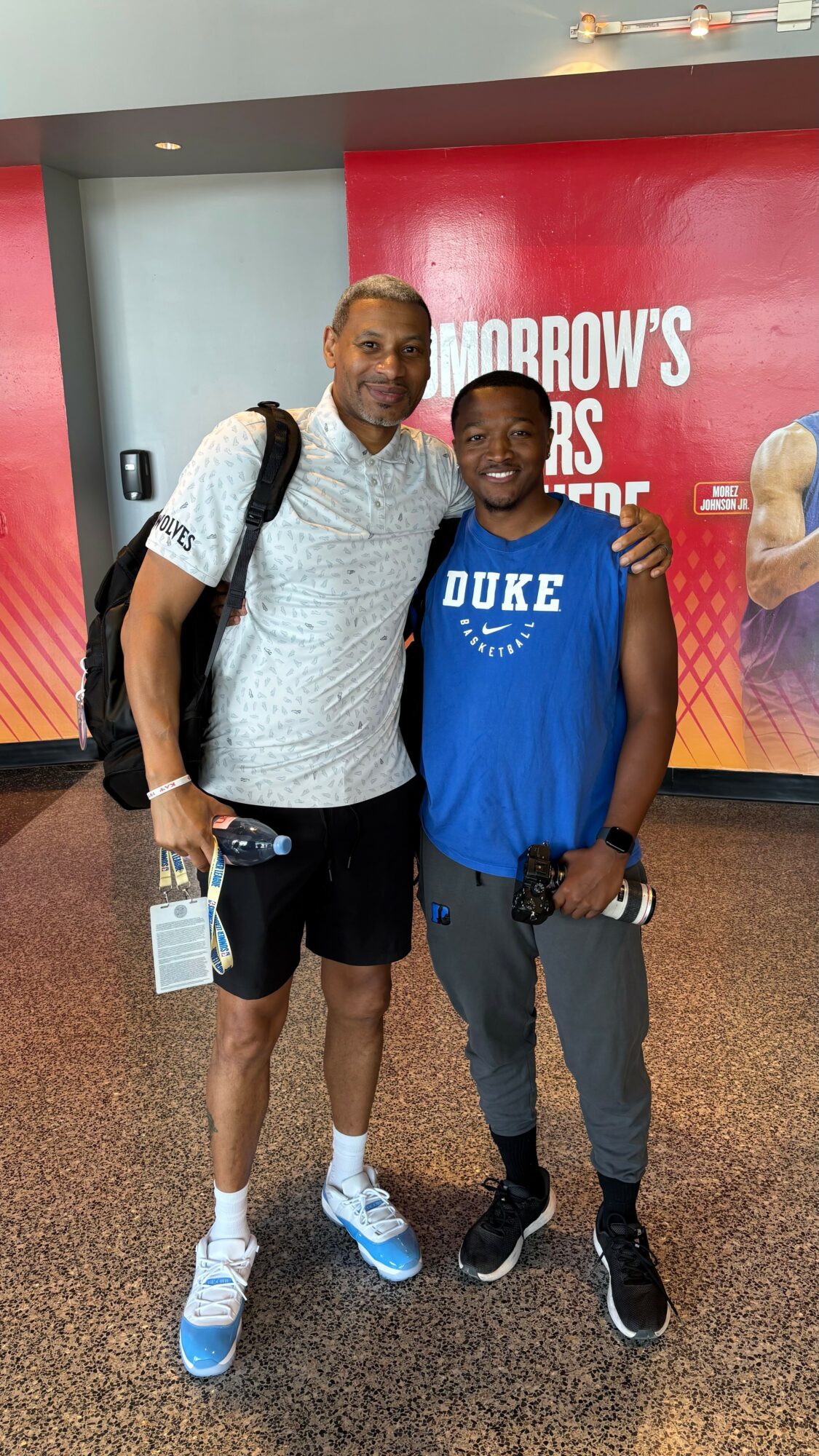 Two people standing together in front of a red and purple background, smiling, one wearing a white shirt and the other a blue Duke shirt.