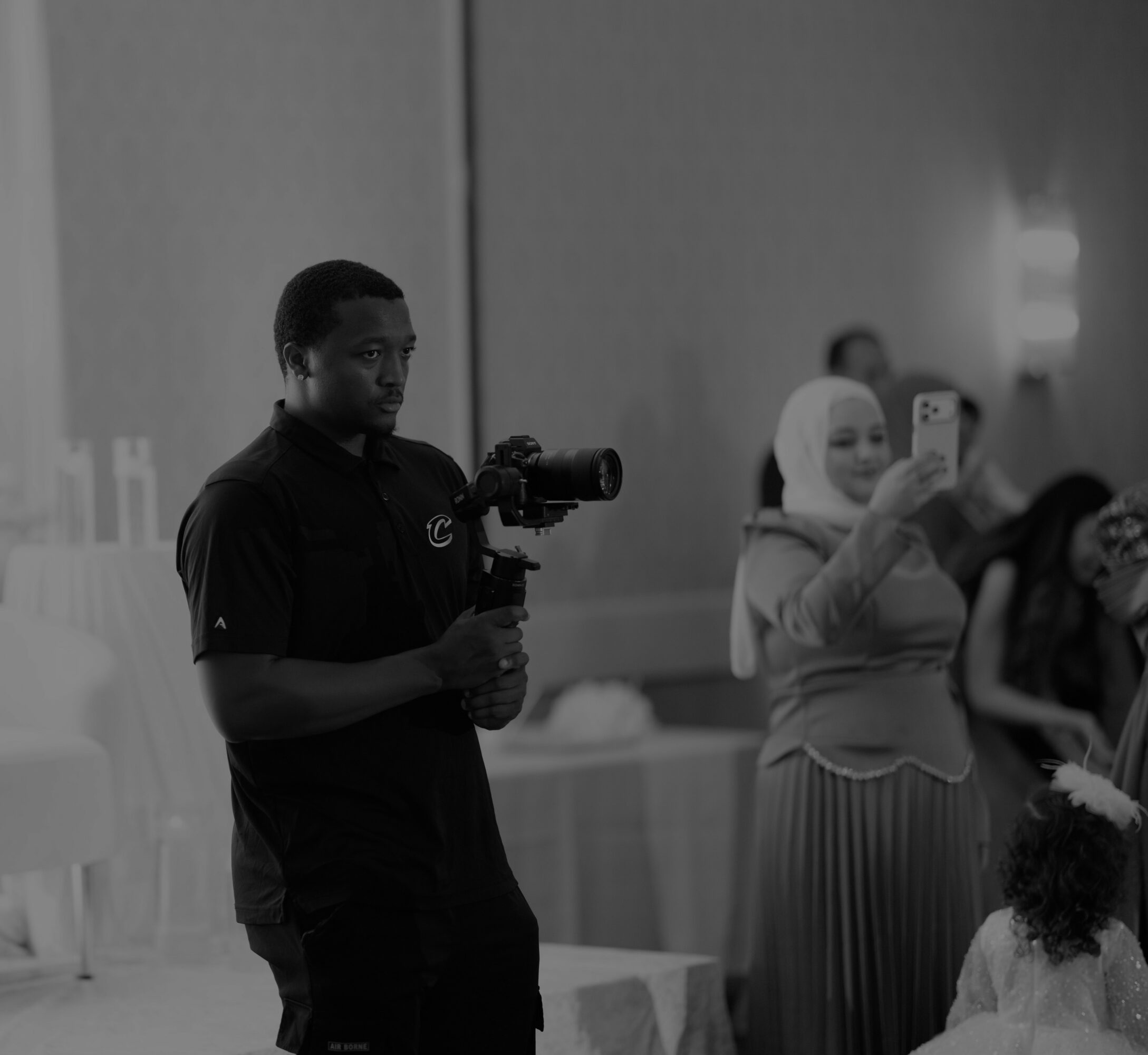 Man holding a camera stabilizer, woman taking a photo with her phone, and a child in a room with a plain wall.