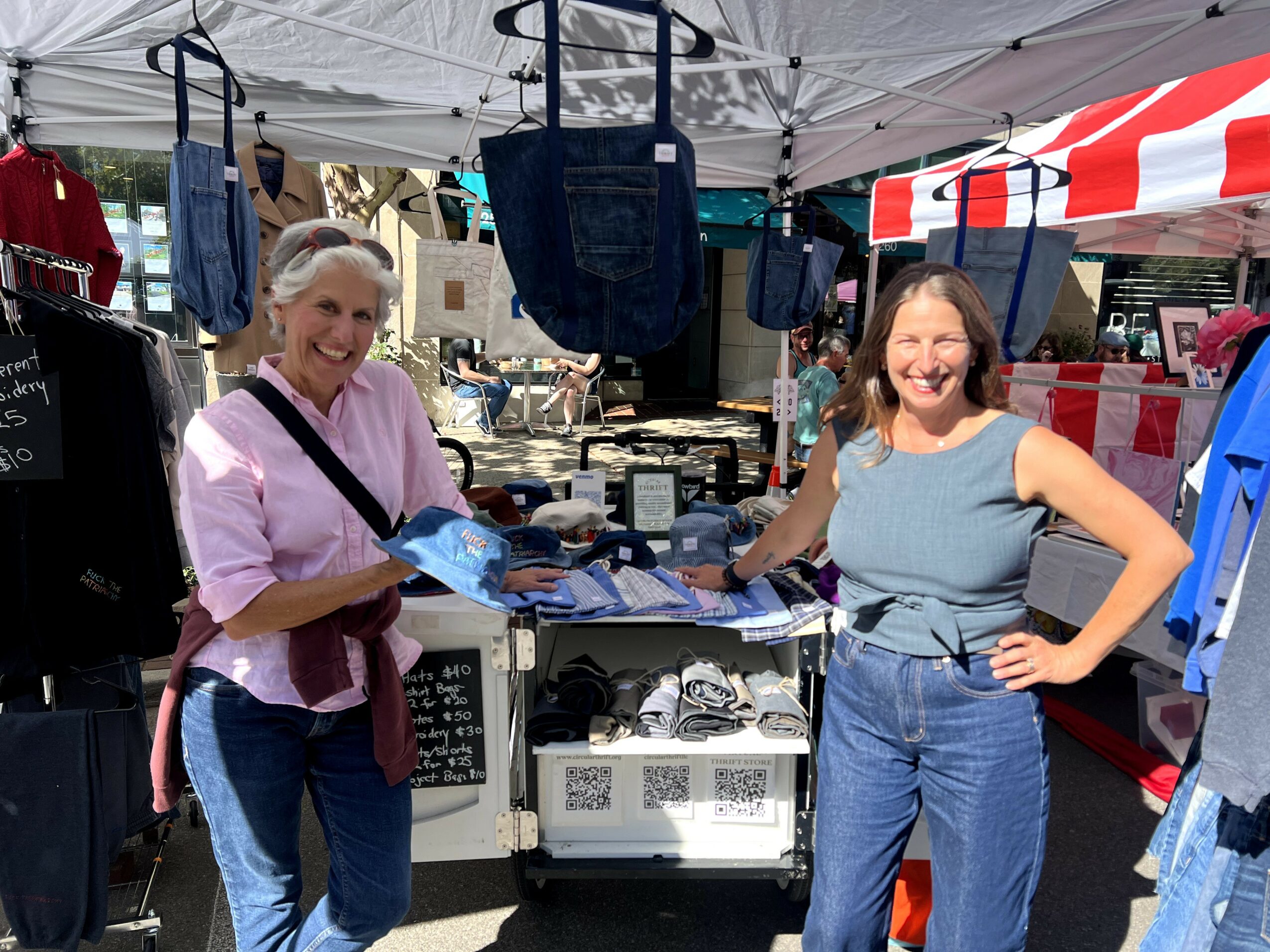 Two women smiling at a market stall with bags and clothing, under a white canopy, in an outdoor setting.