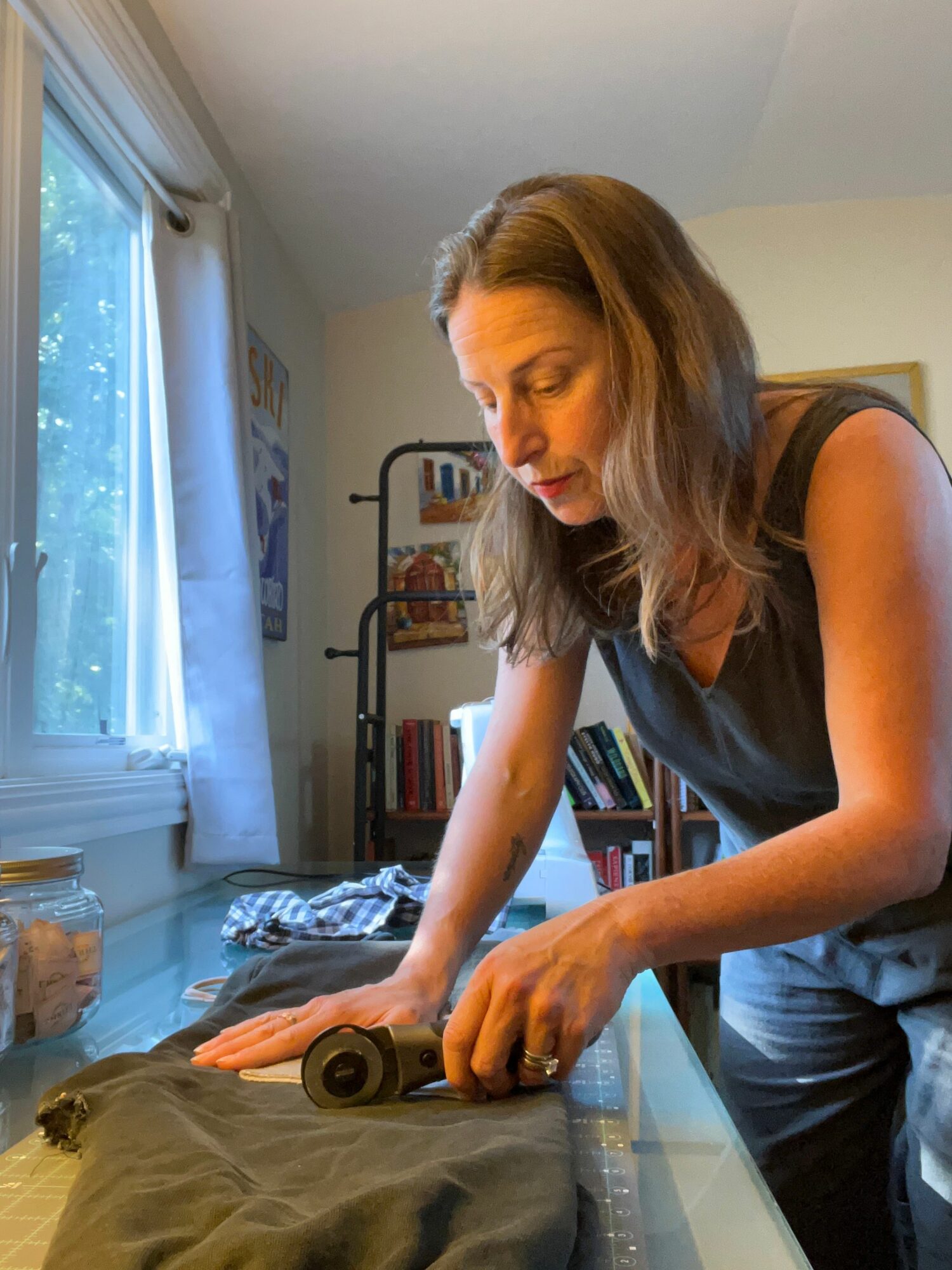 Woman with shoulder-length hair leaning over a table, using a tape measure on a fabric or clothing item, in a room with a window and bookshelf.