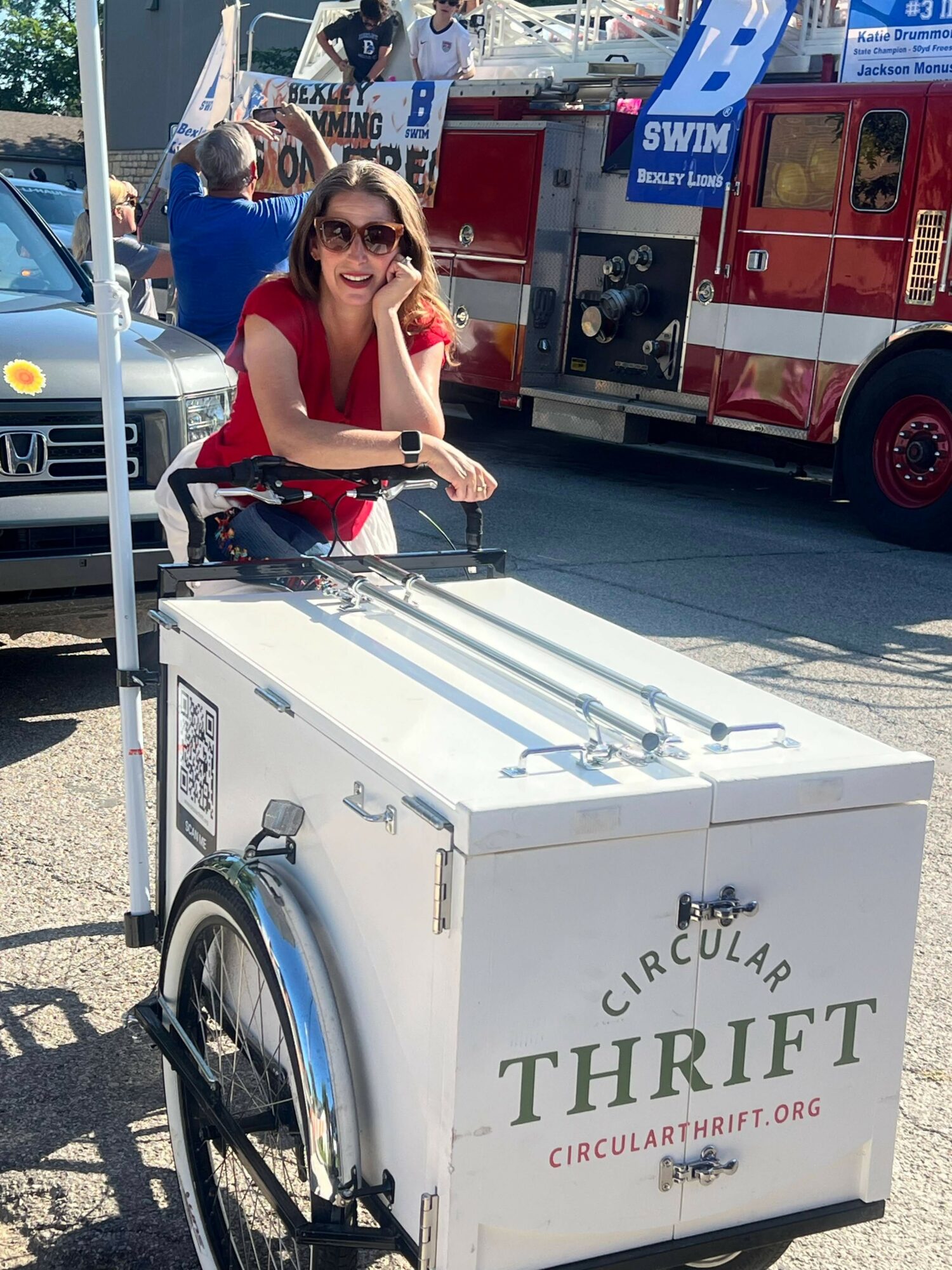 Woman in red dress with sunglasses sitting on a white ice cream cart on a busy city street.