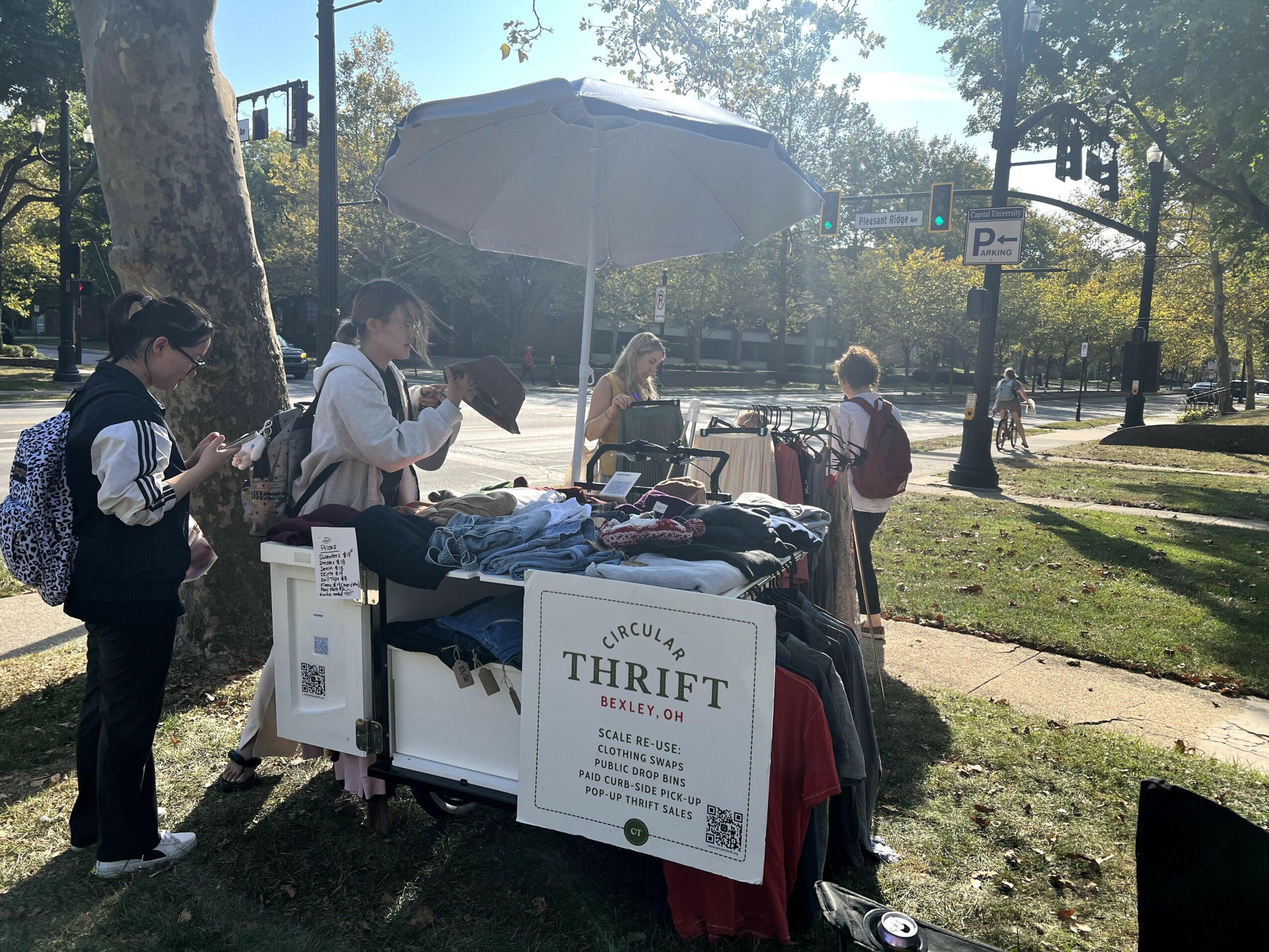 People browsing a thrift sale table outdoors in a park with trees and sunlight, under a large umbrella.