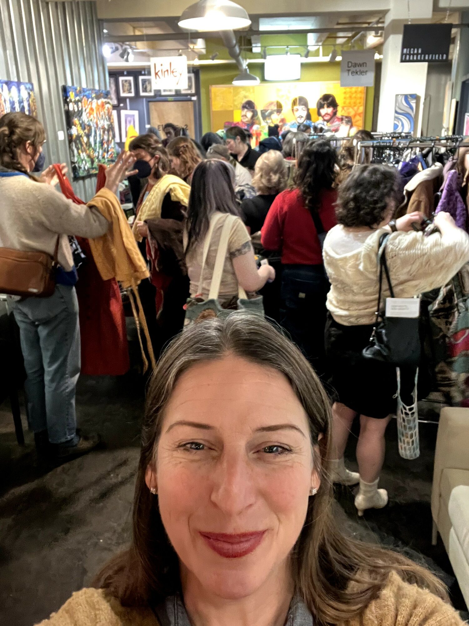 Woman taking selfie in crowded indoor market with people browsing clothing and accessories, colorful displays, and warm lighting.