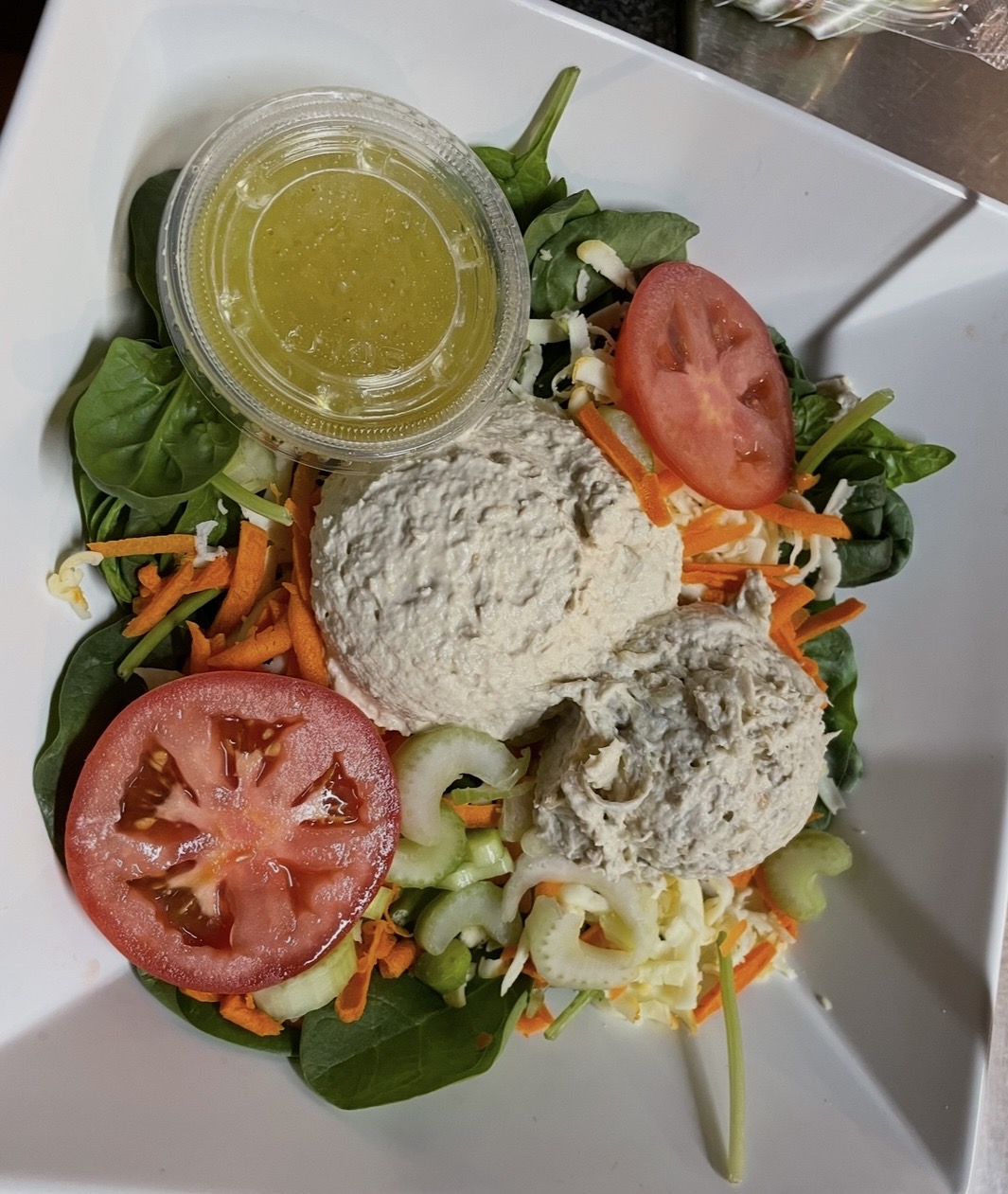 Salad with tomato slices, shredded carrots, greens, two scoops of tuna, and a small container of dressing.