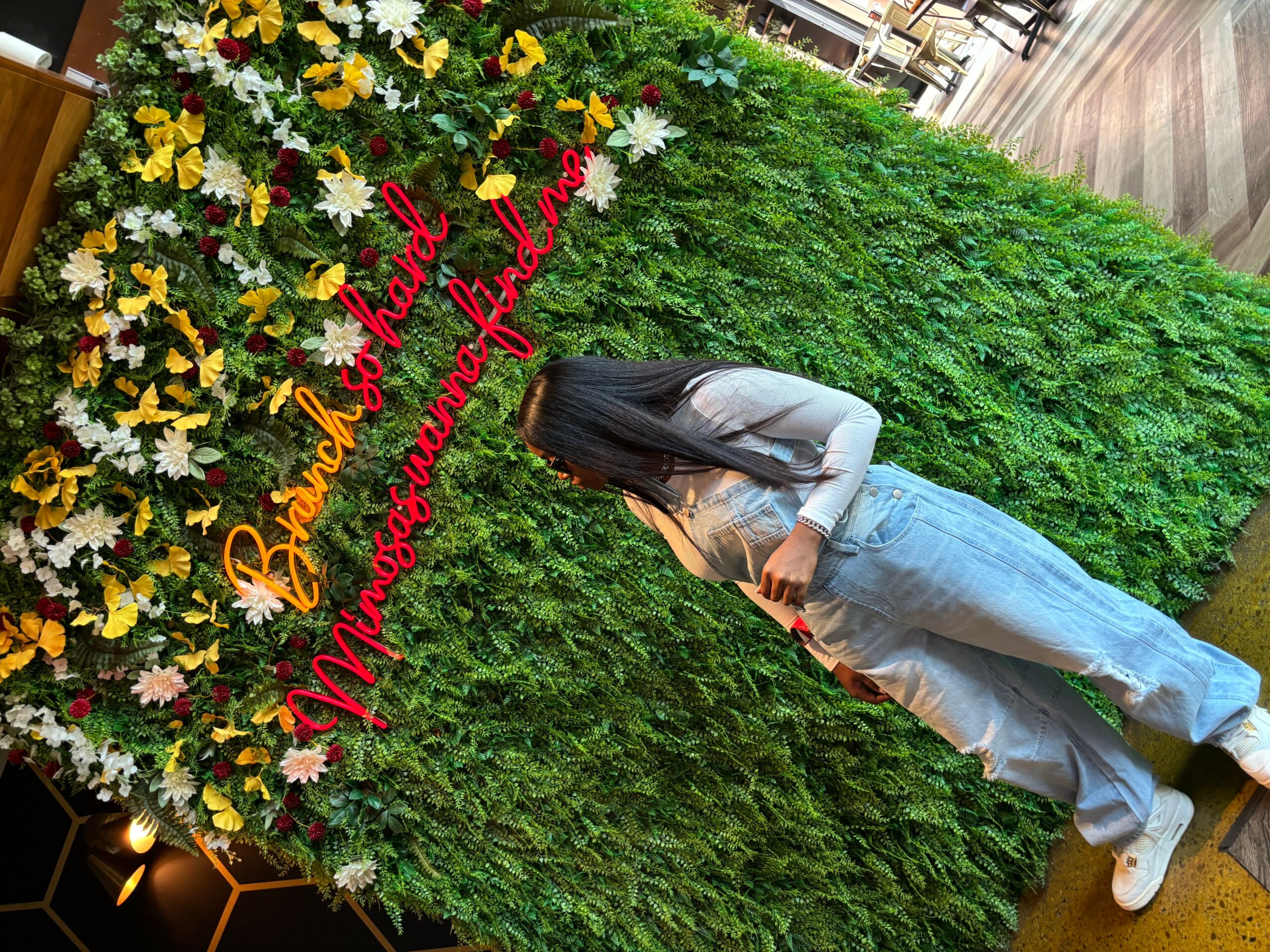 Person with long hair in gray hoodie and jeans standing near a flower wall with a red sign, outdoors.