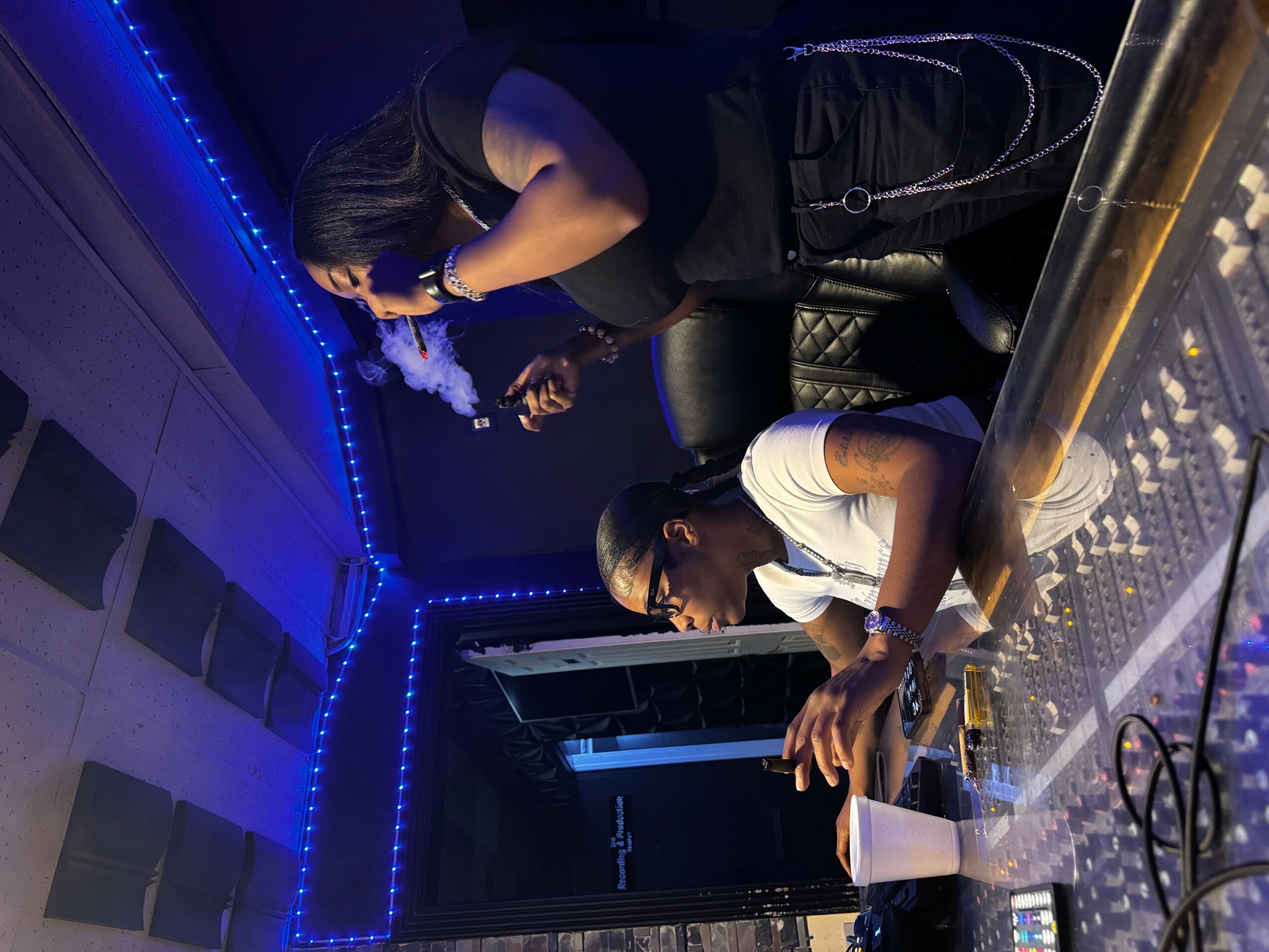 Two women and a man at a bar counter, one woman leaning over, the other sitting, with blue lighting in the background.