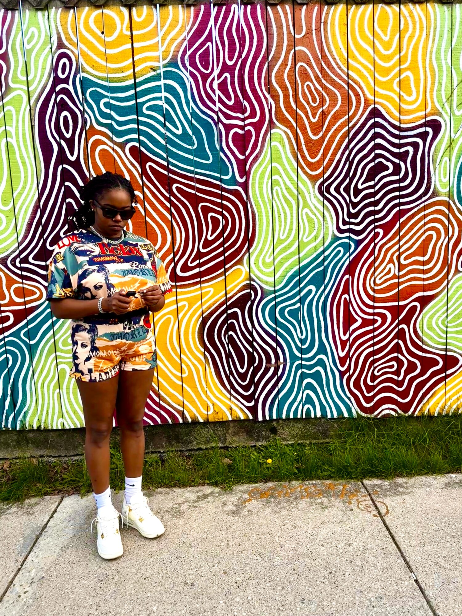Girl in colorful outfit and sunglasses standing in front of a vibrant, abstract mural wall.