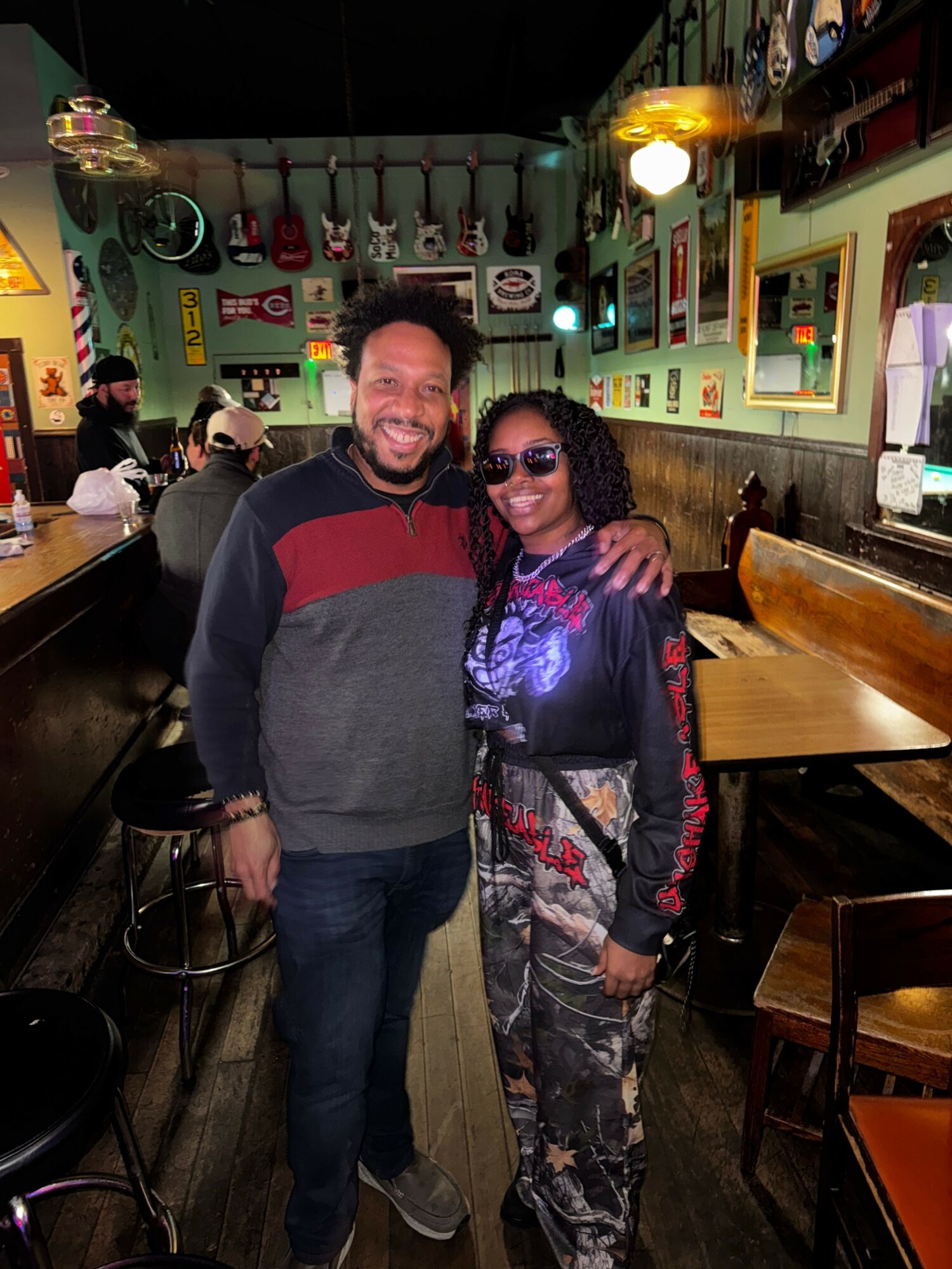 Two people standing in a bar or restaurant, smiling, with colorful decor and tables in the background.