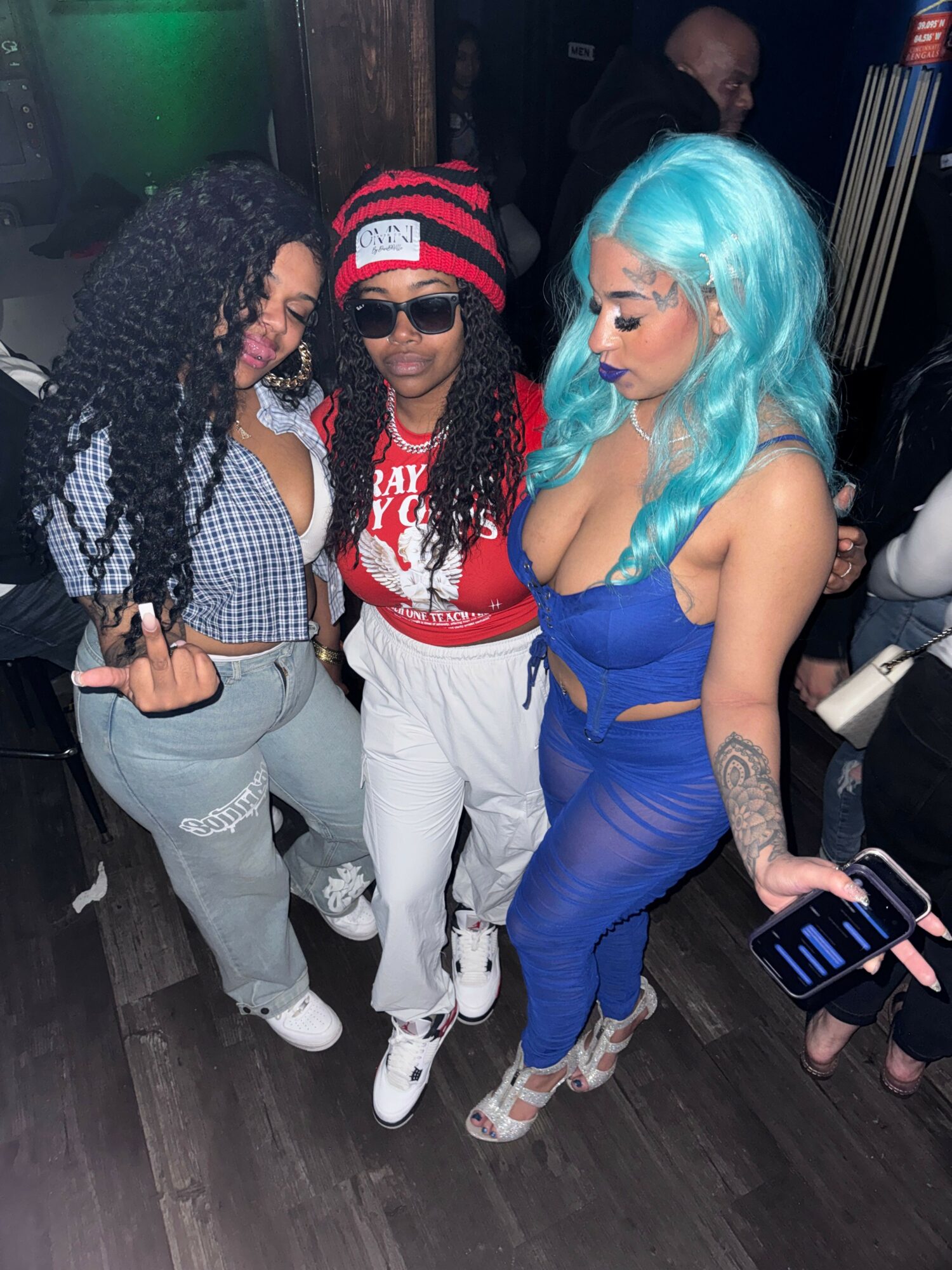 Three people with colorful hair and casual clothing standing together in a dark indoor setting.