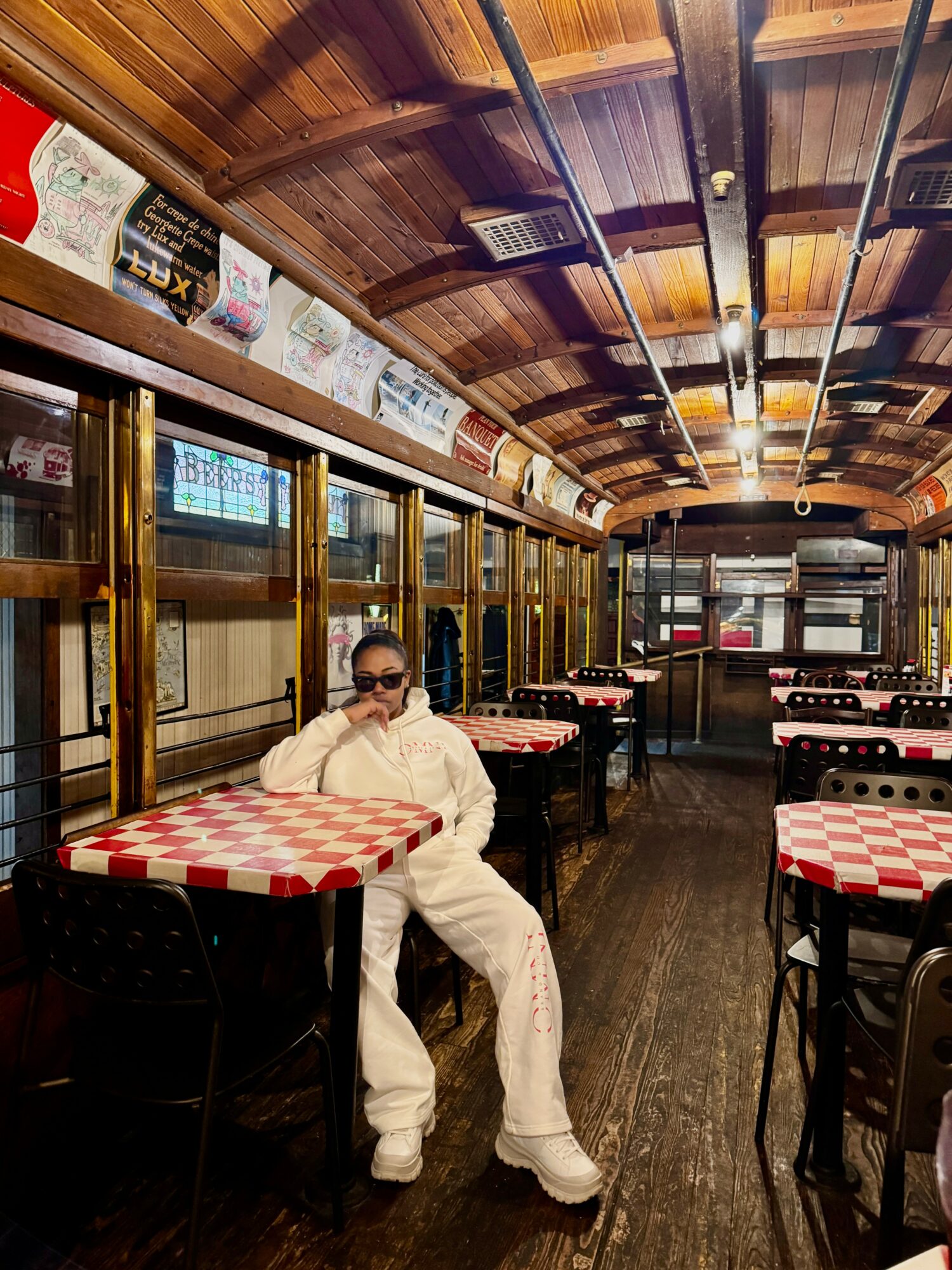 Person sitting at a table in a restaurant with wooden ceiling and large windows, wearing sunglasses and white outfit.