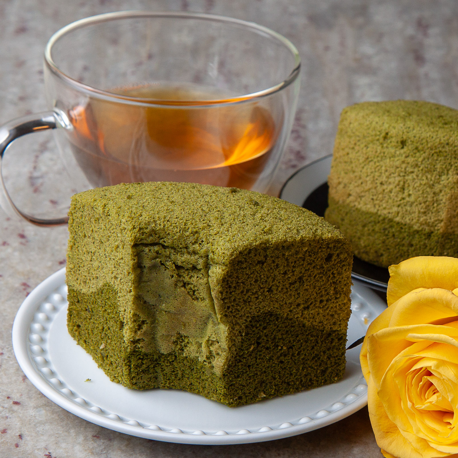 A slice of green tea cake with filling on a white plate, a whole cake behind, a yellow rose, and a glass of tea.