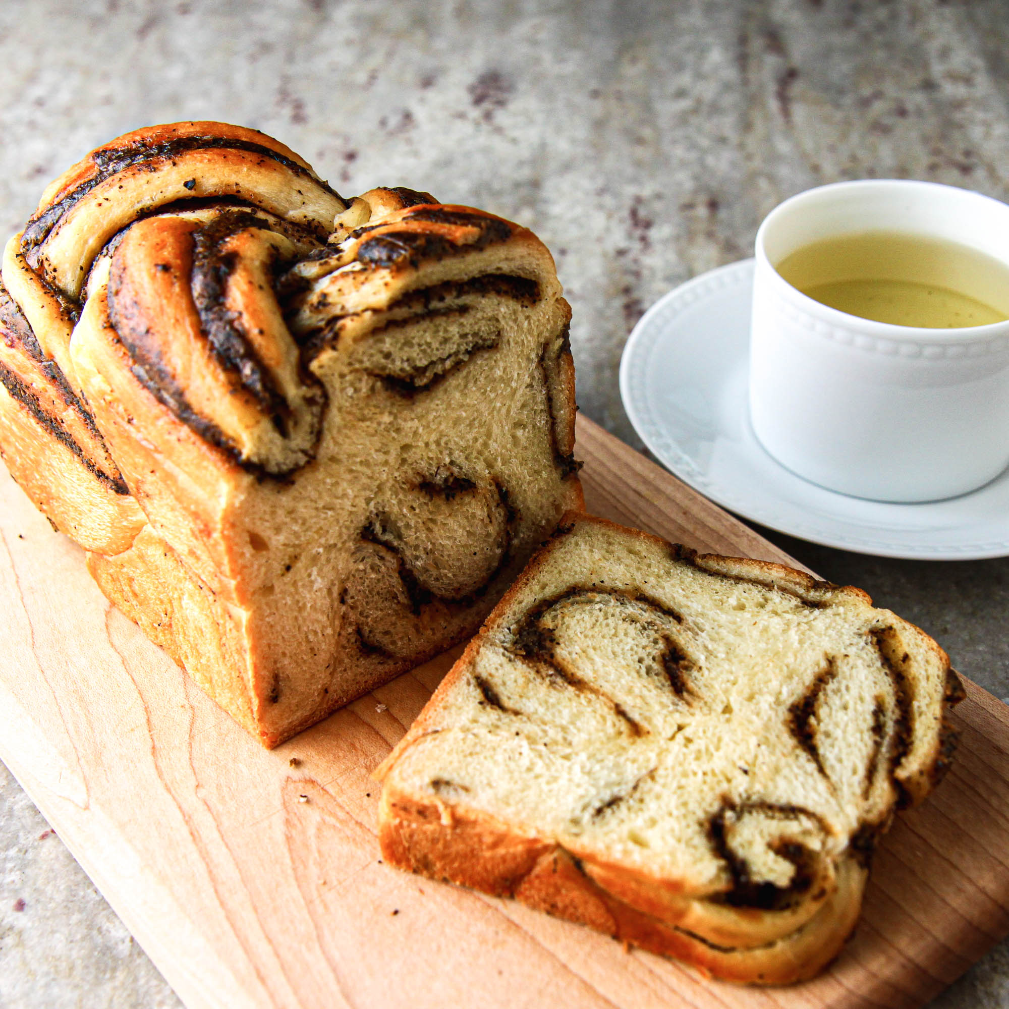Loaf of marble swirl bread with a slice cut, placed on a wooden board with a cup of tea or coffee nearby.