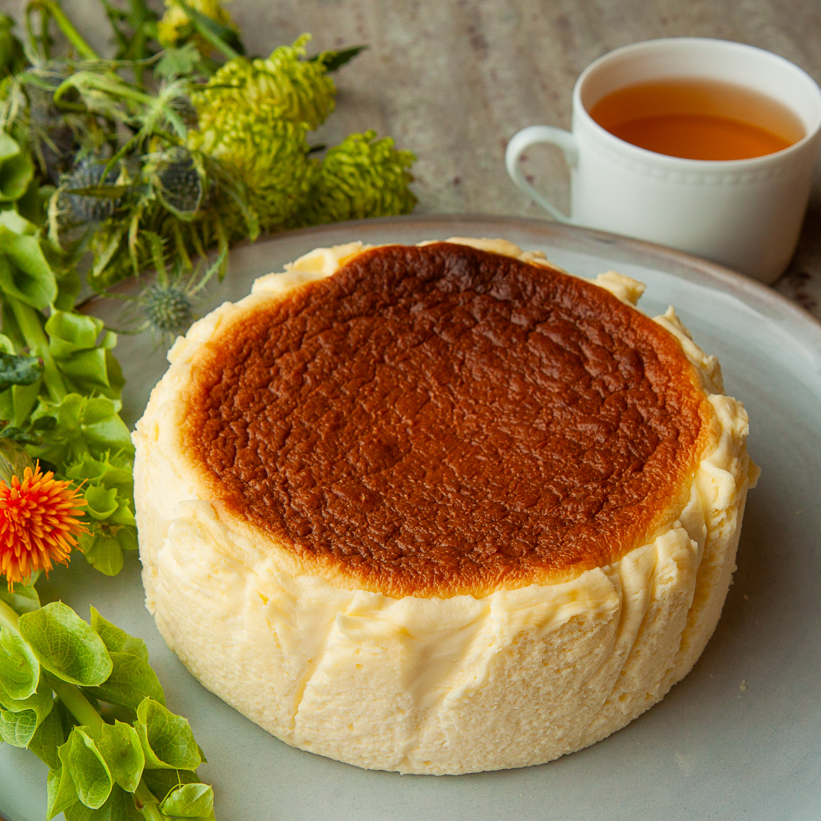 Cheesecake with a golden-brown top on a plate, surrounded by green plants and a cup of tea in the background.