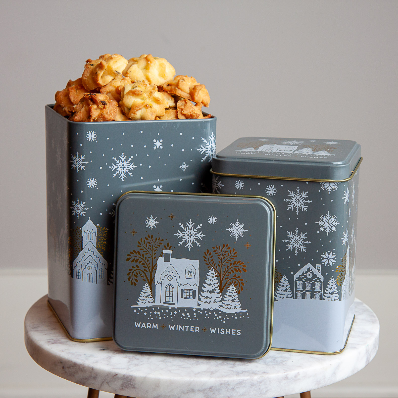 Three metal tins with winter village designs, filled with popcorn, on a white marble surface against a plain background.