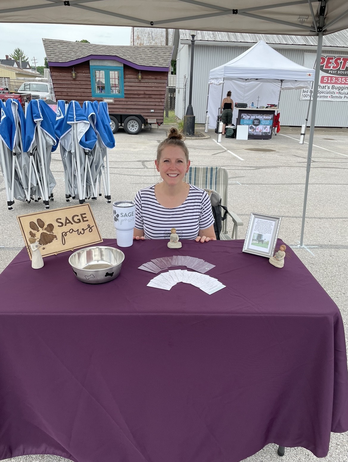 Young woman sitting at a table with a purple tablecloth, smiling, with flyers and a framed photo, outdoors at a fair.