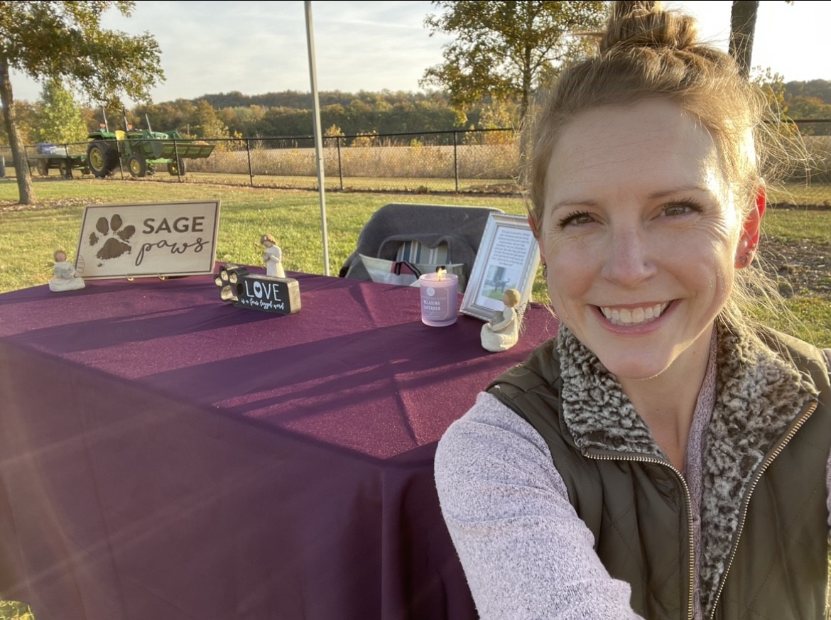 Smiling woman taking selfie outdoors with a table displaying signs and items, trees, and a field in the background.