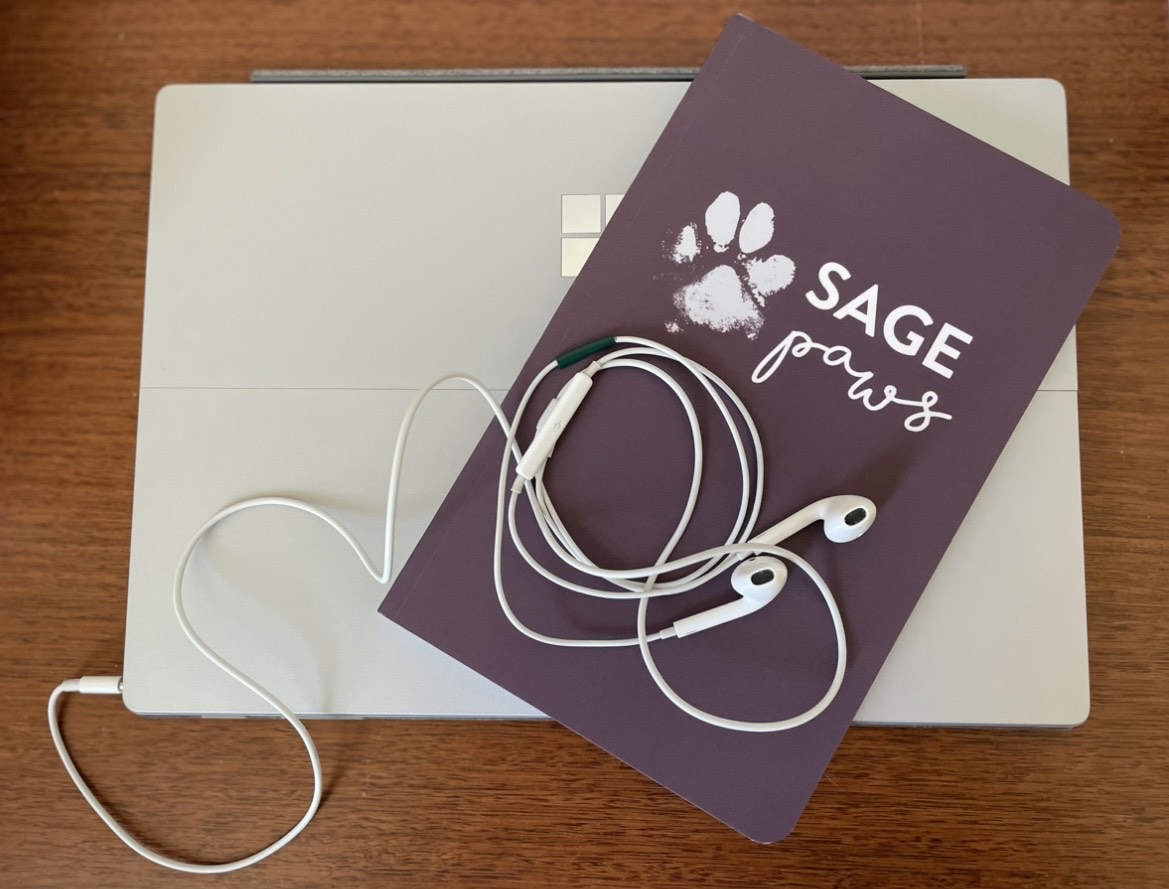 Laptop, purple booklet titled 'SAGE Paws,' and white earphones on a wooden surface.