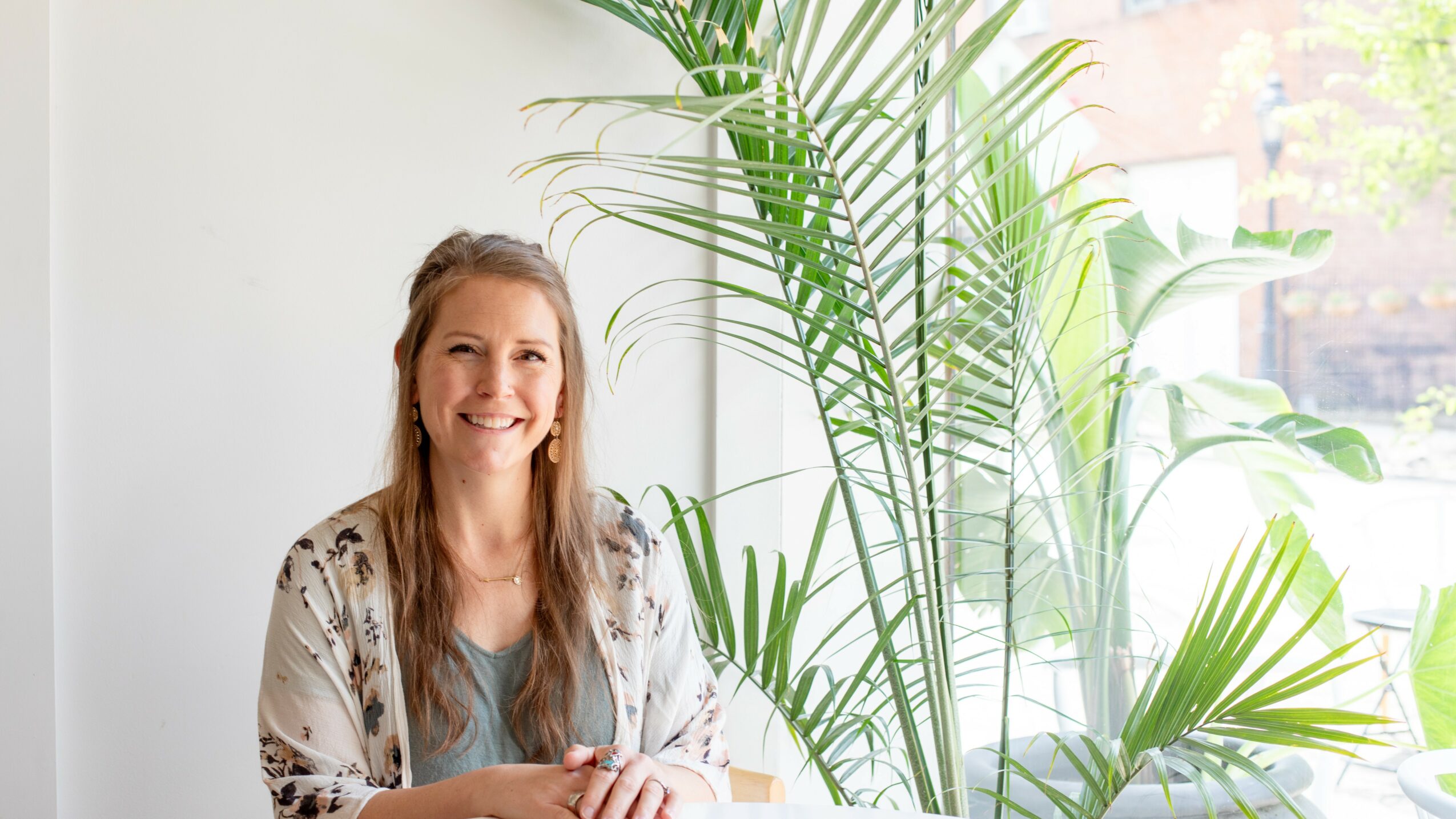 Woman with long hair smiling, sitting near large green plants and a window, bright natural light, casual clothing