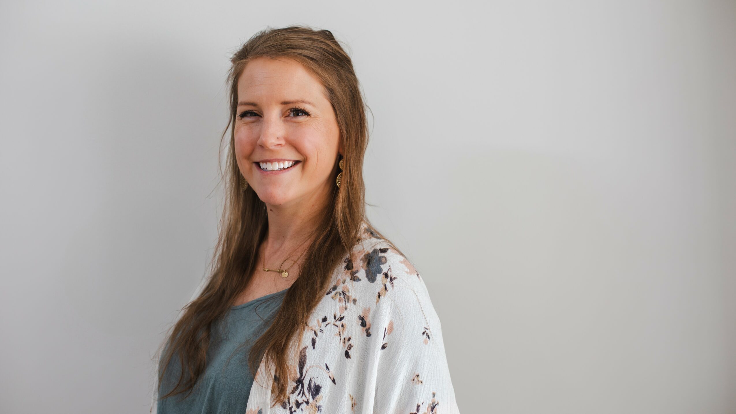 Smiling woman with long hair wearing a floral cardigan and blue top, standing against a plain light background.