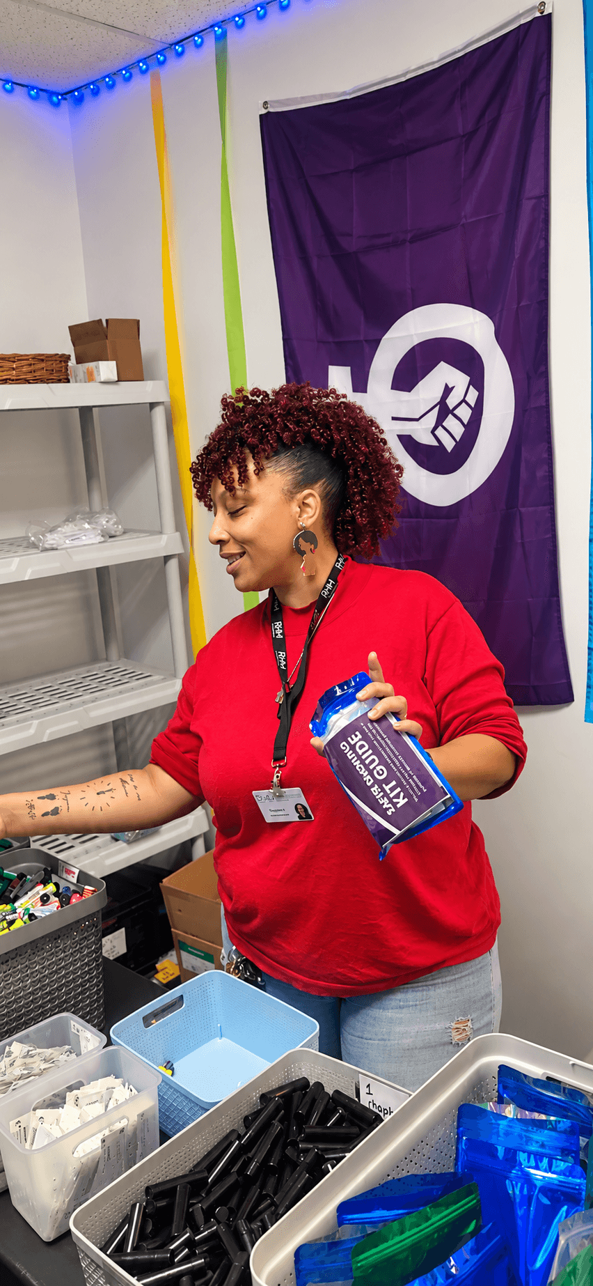Woman with curly hair in red shirt holding a box, standing near shelves with supplies, purple flag on wall behind her.