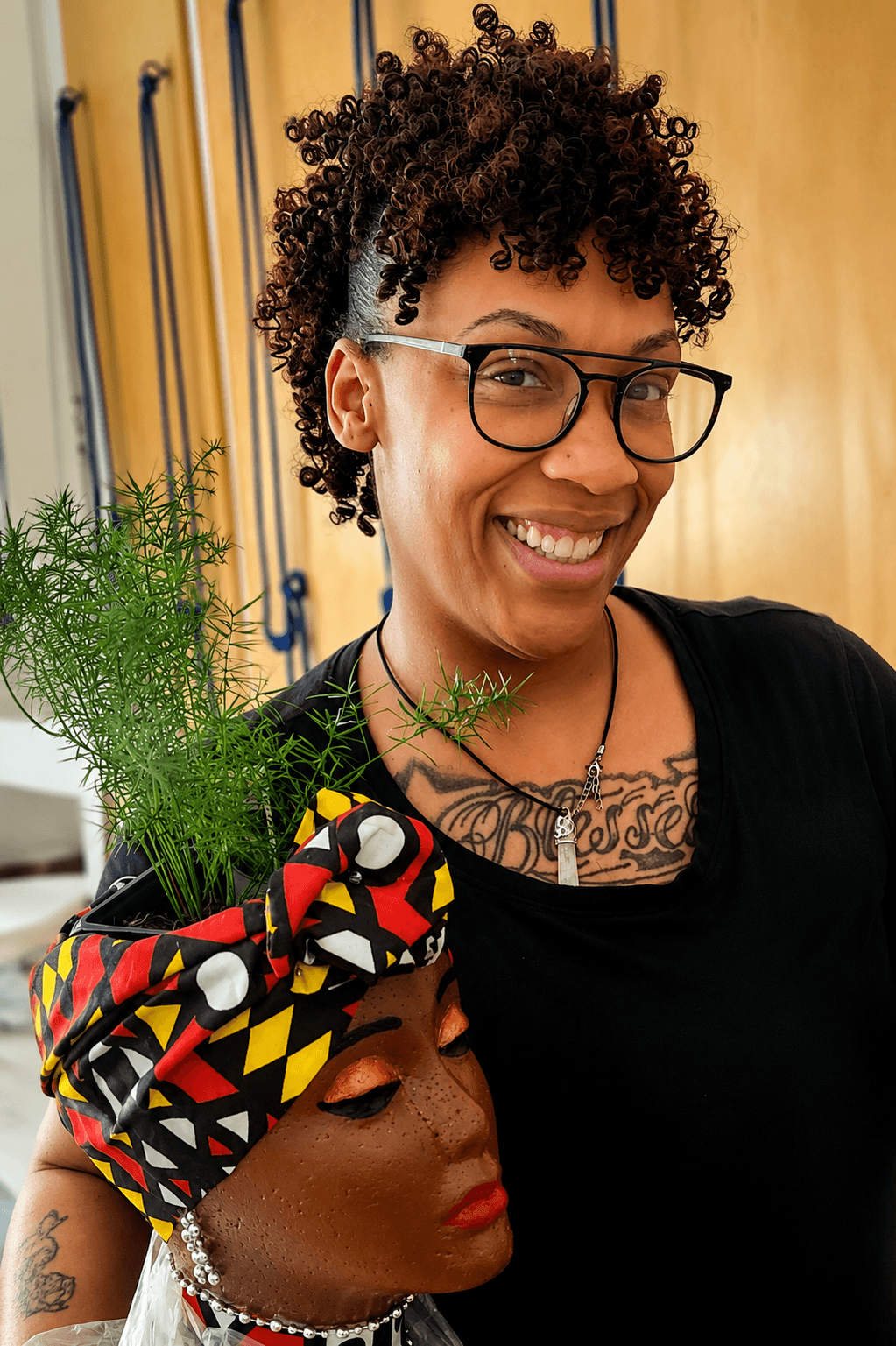 Smiling woman with curly hair and glasses, wearing a black top, holding a plant, with colorful headwrap and jewelry.
