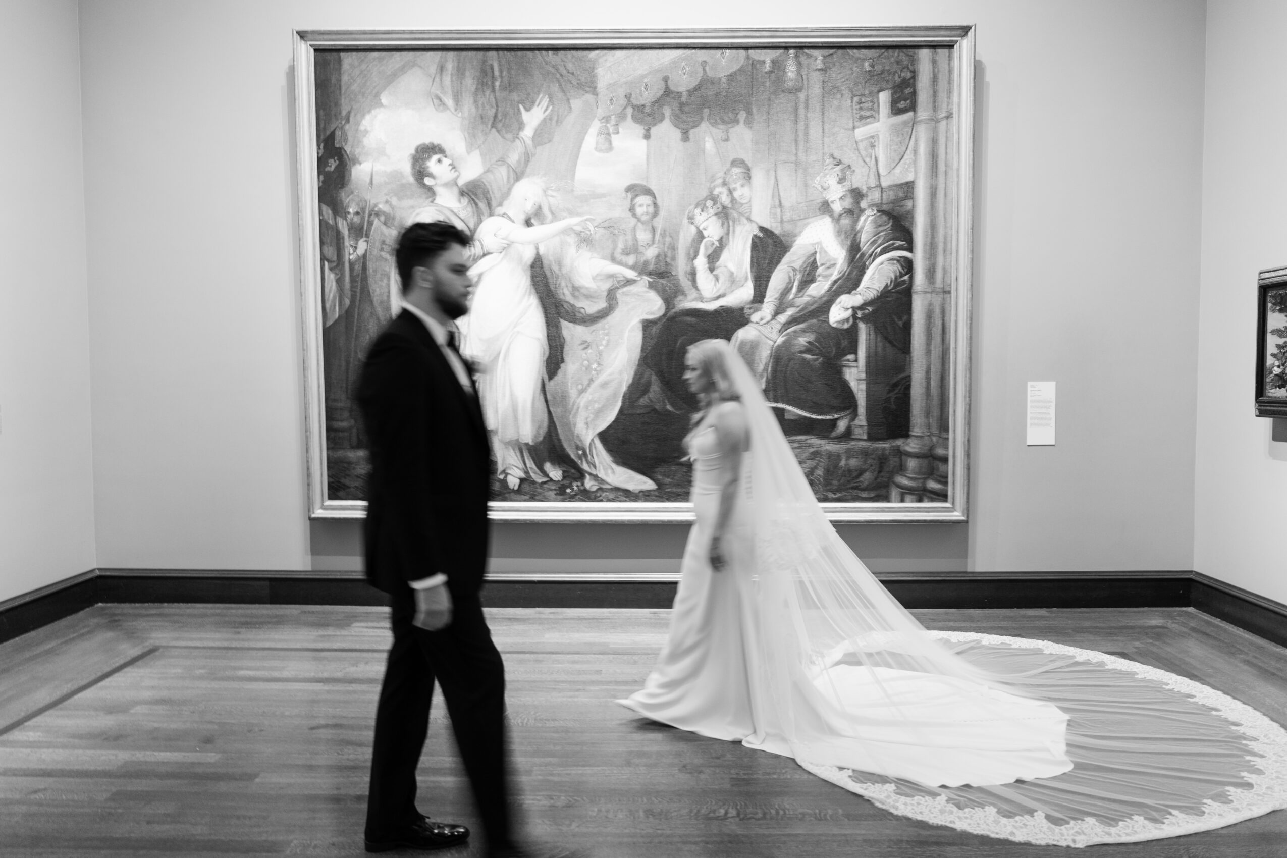 A bride and groom stand in an art gallery, with a large painting behind them, the bride in a white gown and the groom in a suit.