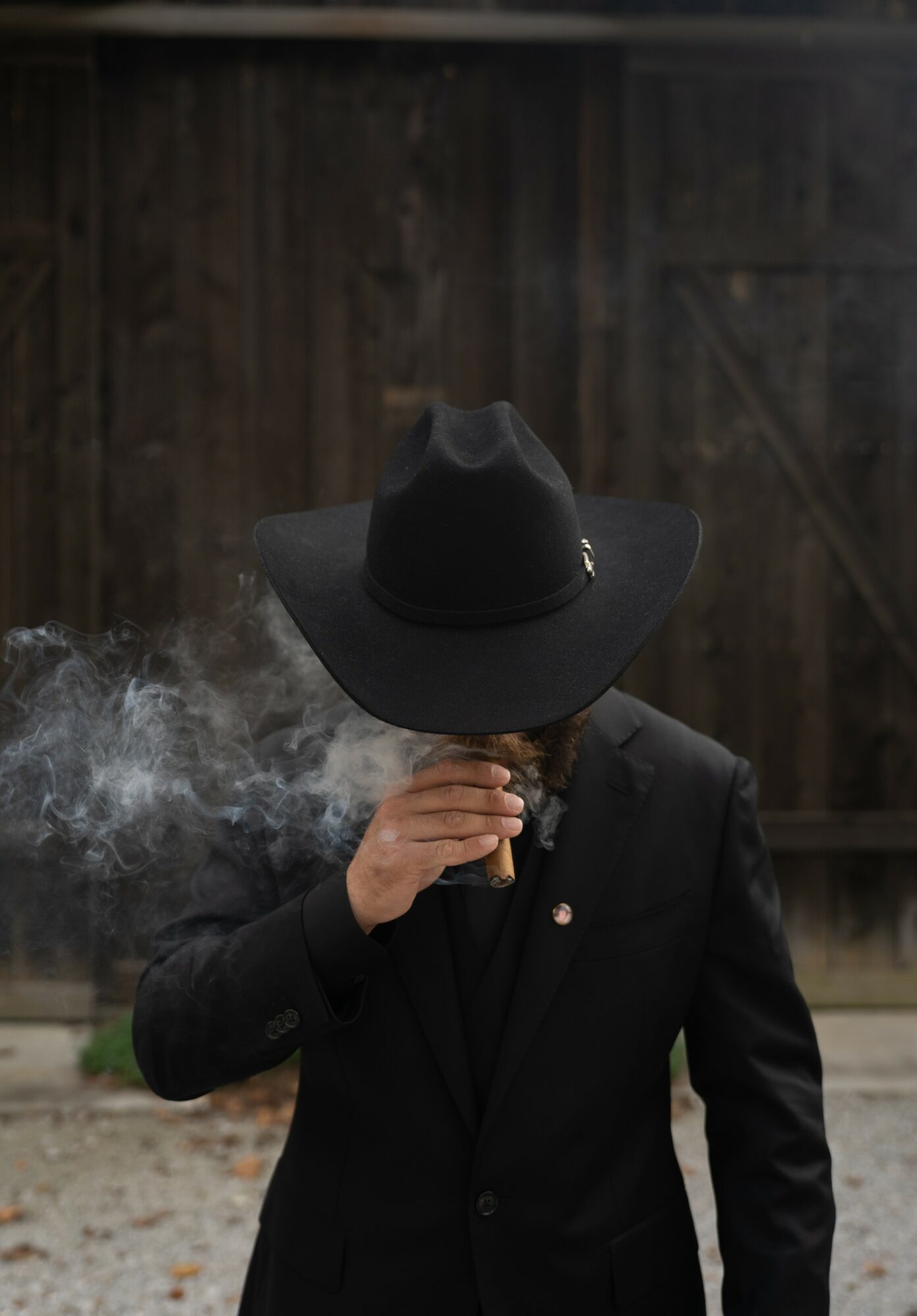 Person wearing a large black hat and black suit, smoking a cigarette, with a wooden fence background.