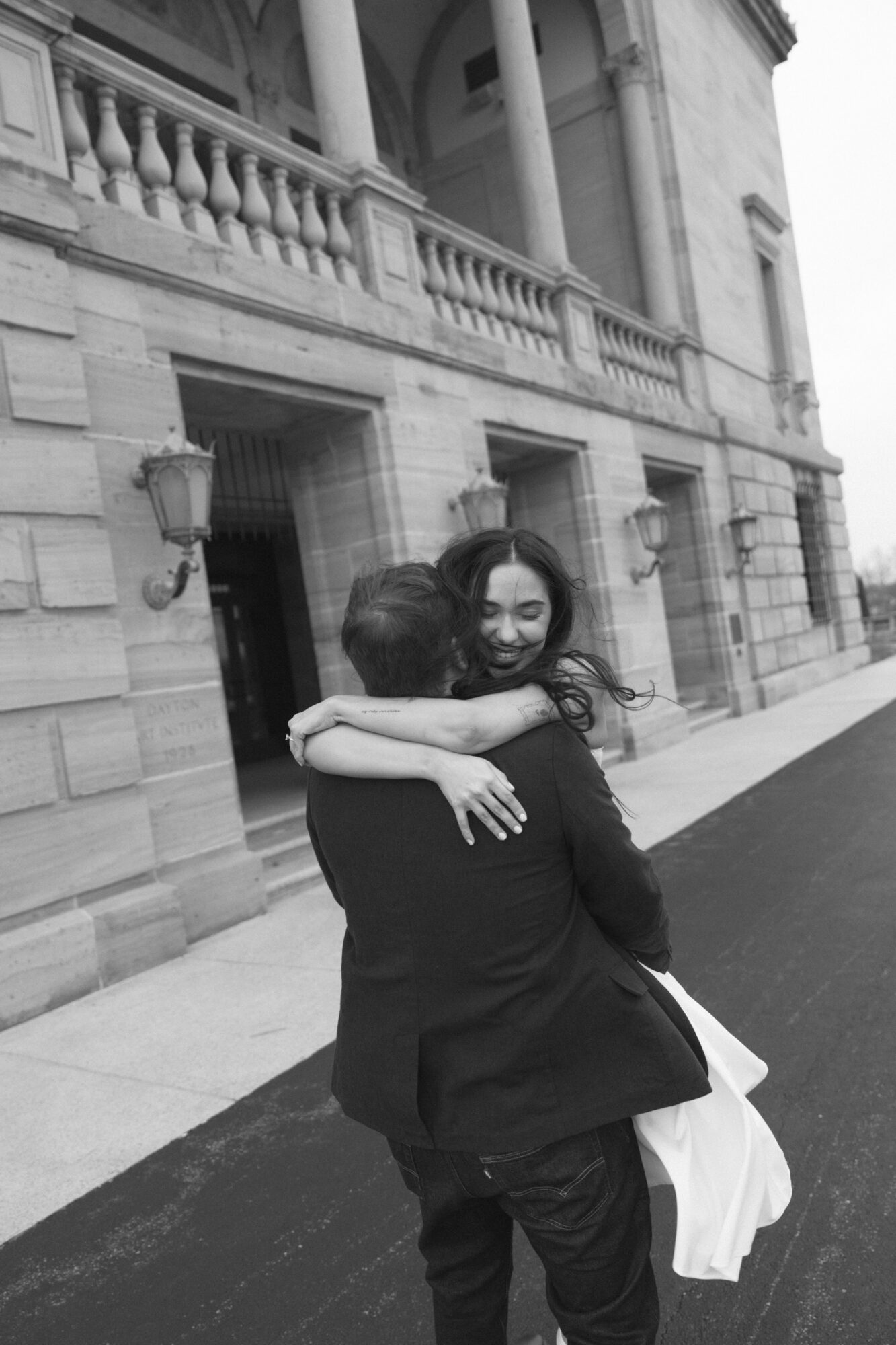 Two people hugging outside a building with classical architecture, one person's face visible, the other with back to camera.