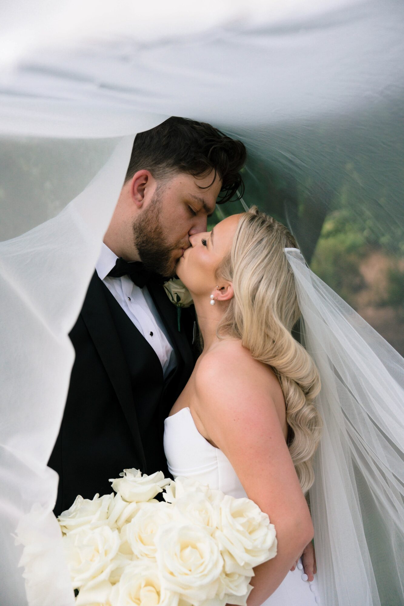 Bride and groom share a kiss under a transparent canopy, bride holding a bouquet of white roses, wedding attire visible.