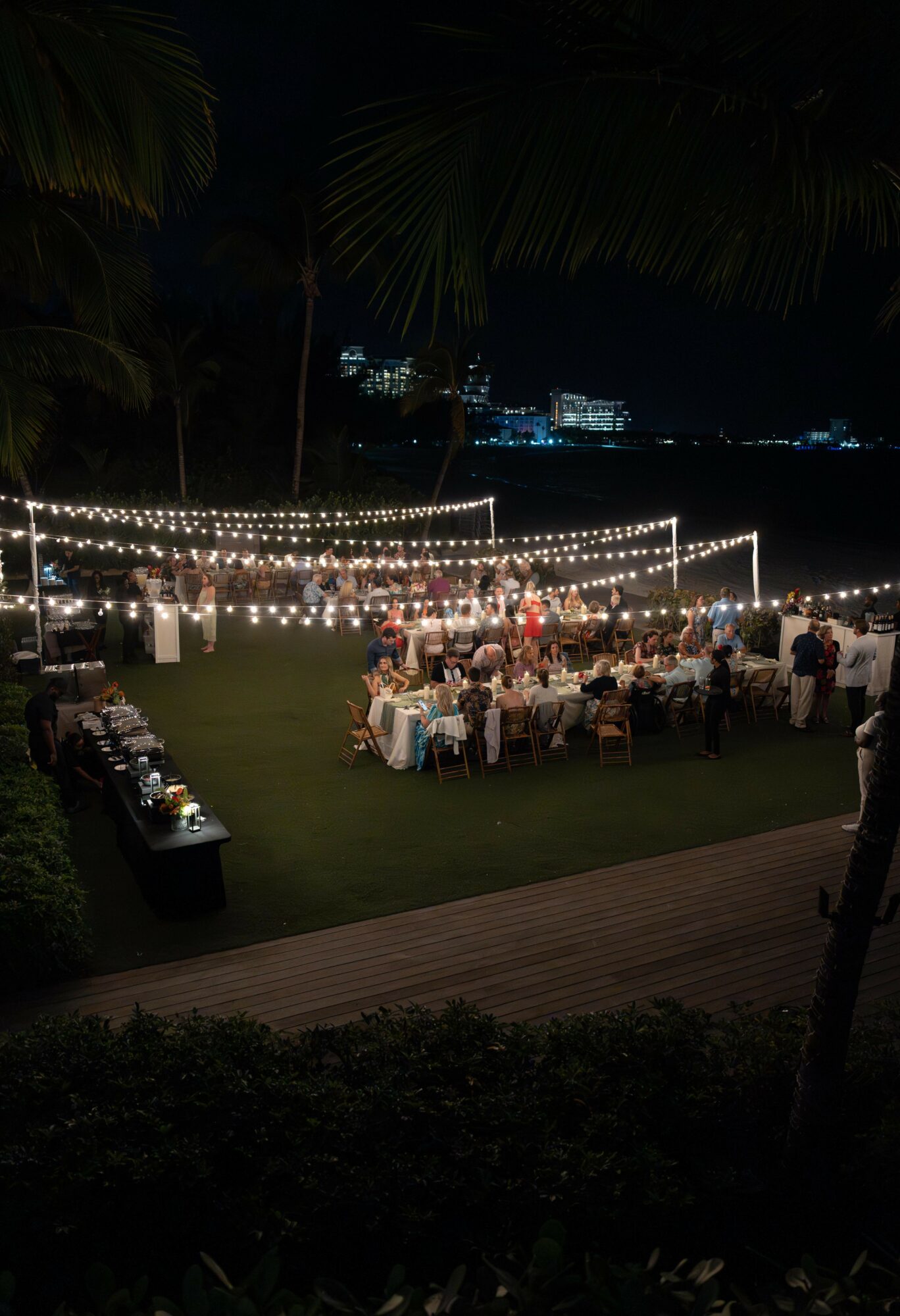 Night outdoor event with string lights, tables, and people, under trees with city lights in background.