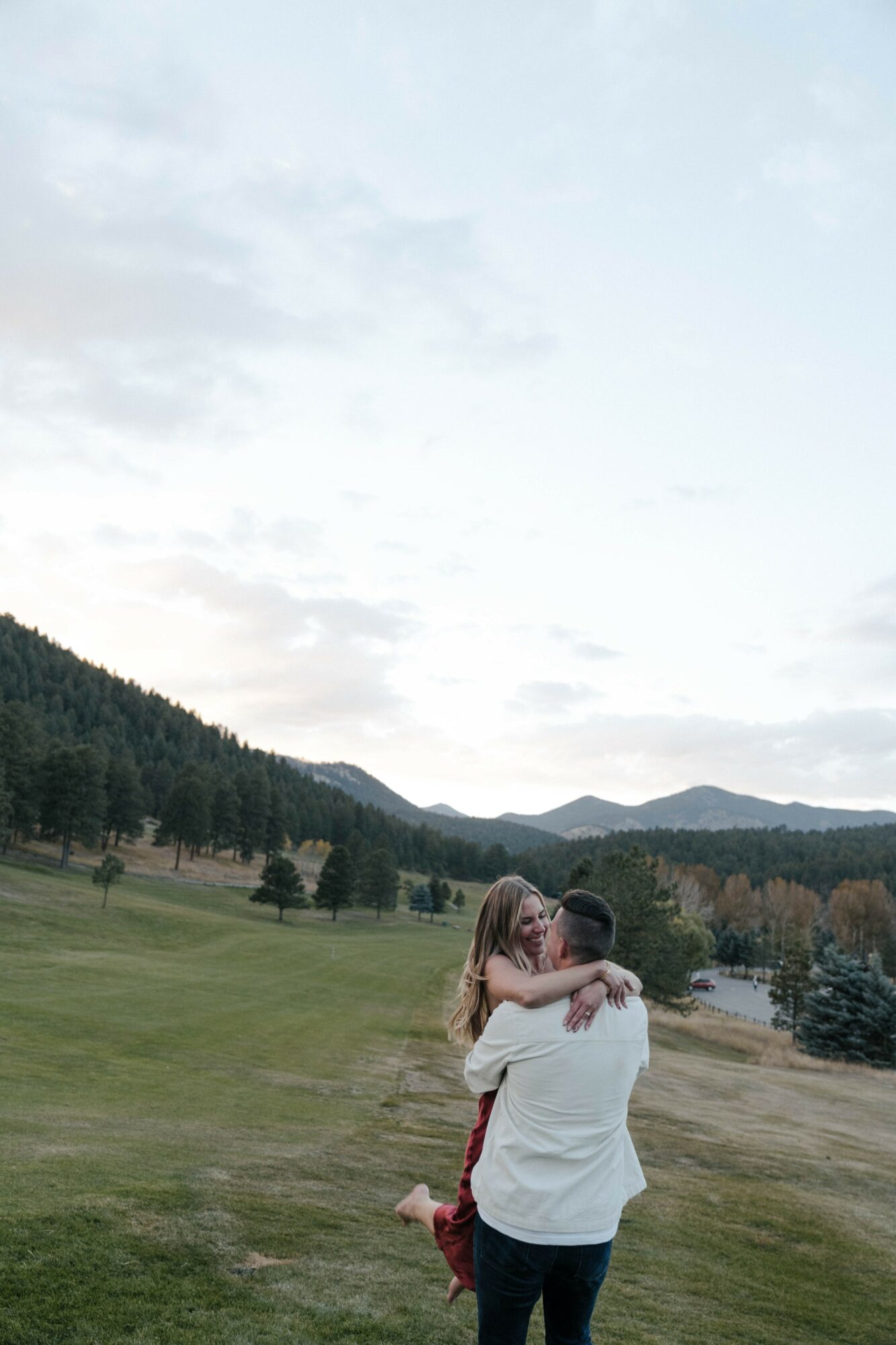 Two people hugging outdoors with a landscape of trees, hills, and a cloudy sky in the background.