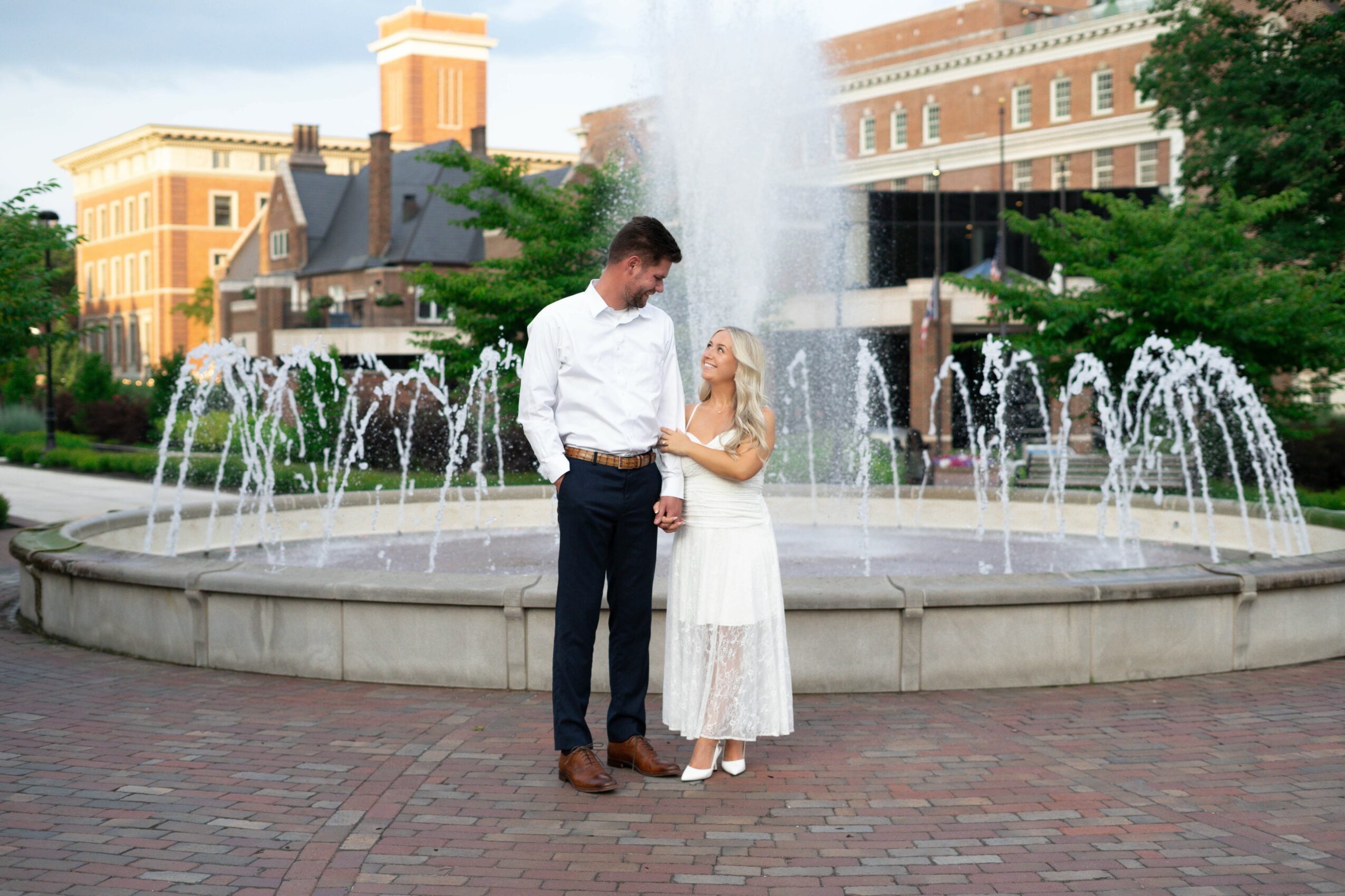 A man and woman stand near a fountain with water jets, holding hands and looking at each other, in an urban park setting.