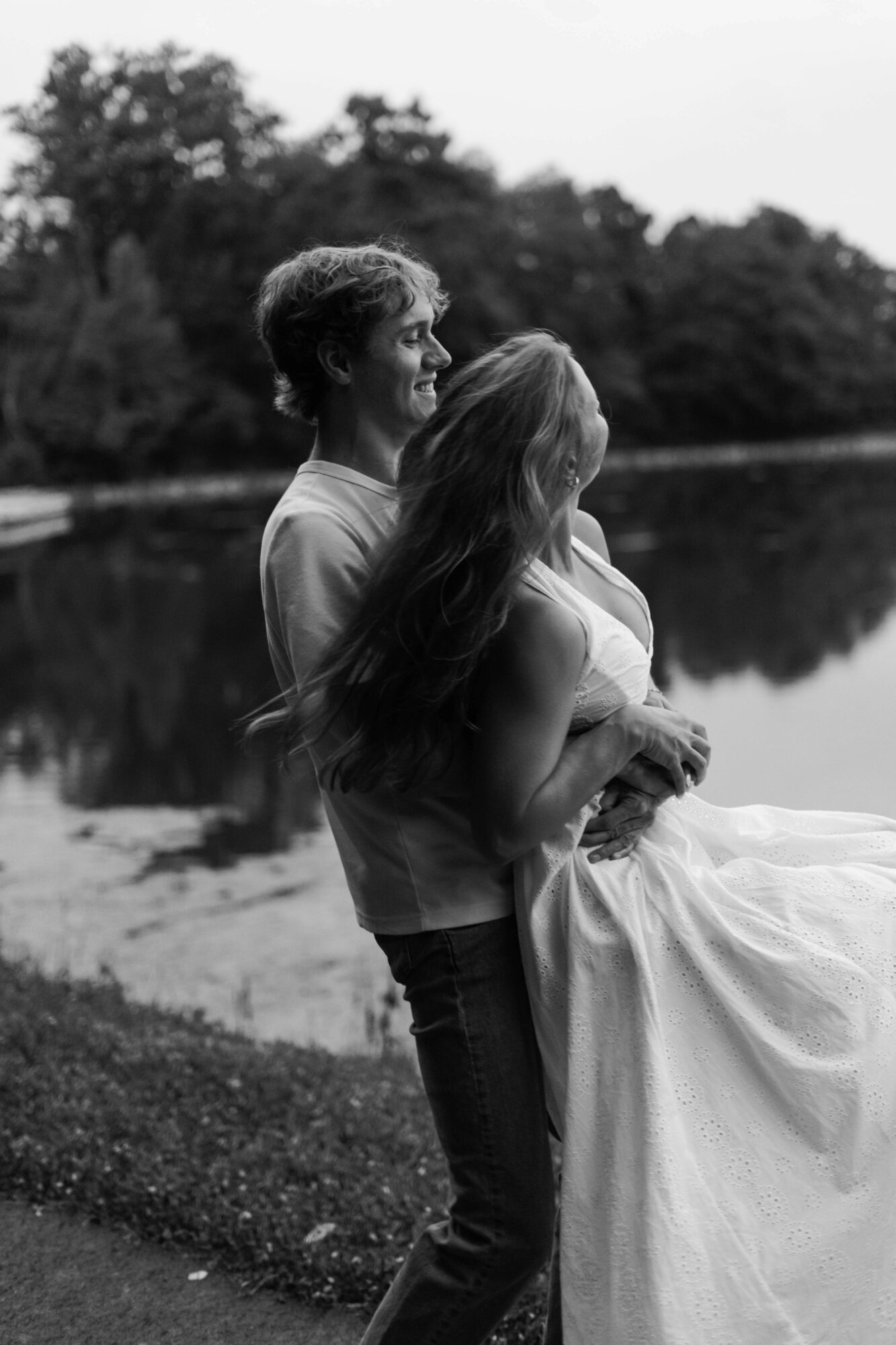A couple stands near a body of water, with trees in the background, the woman in a dress and the man smiling.
