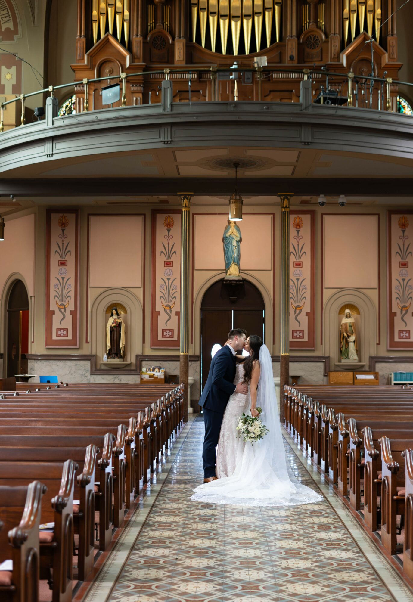 Bride and groom kiss in church aisle with pews and religious statues, ornate interior, and organ pipes above, in a wedding scene.