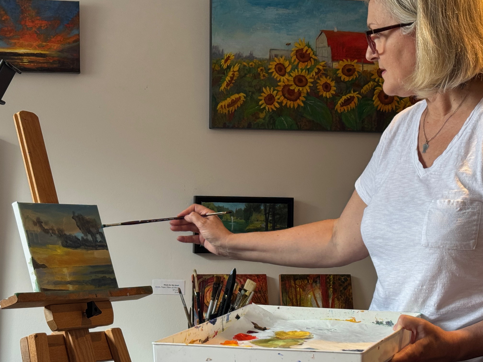 Woman painting on canvas in art studio with colorful paintings on wall, holding a paintbrush, palette in hand.