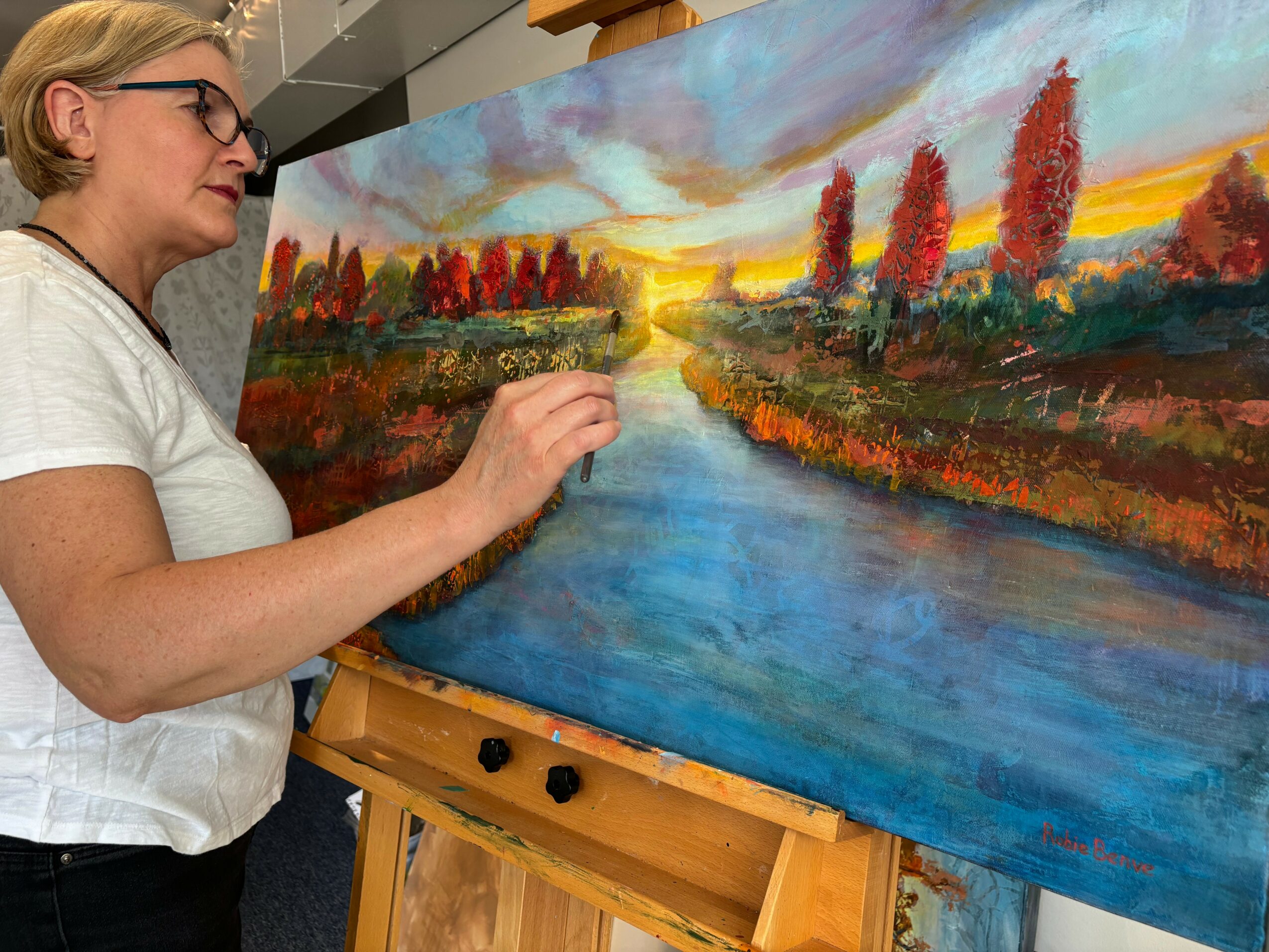 Woman painting a landscape with trees, river, and sunset on a large canvas, standing in an art studio.