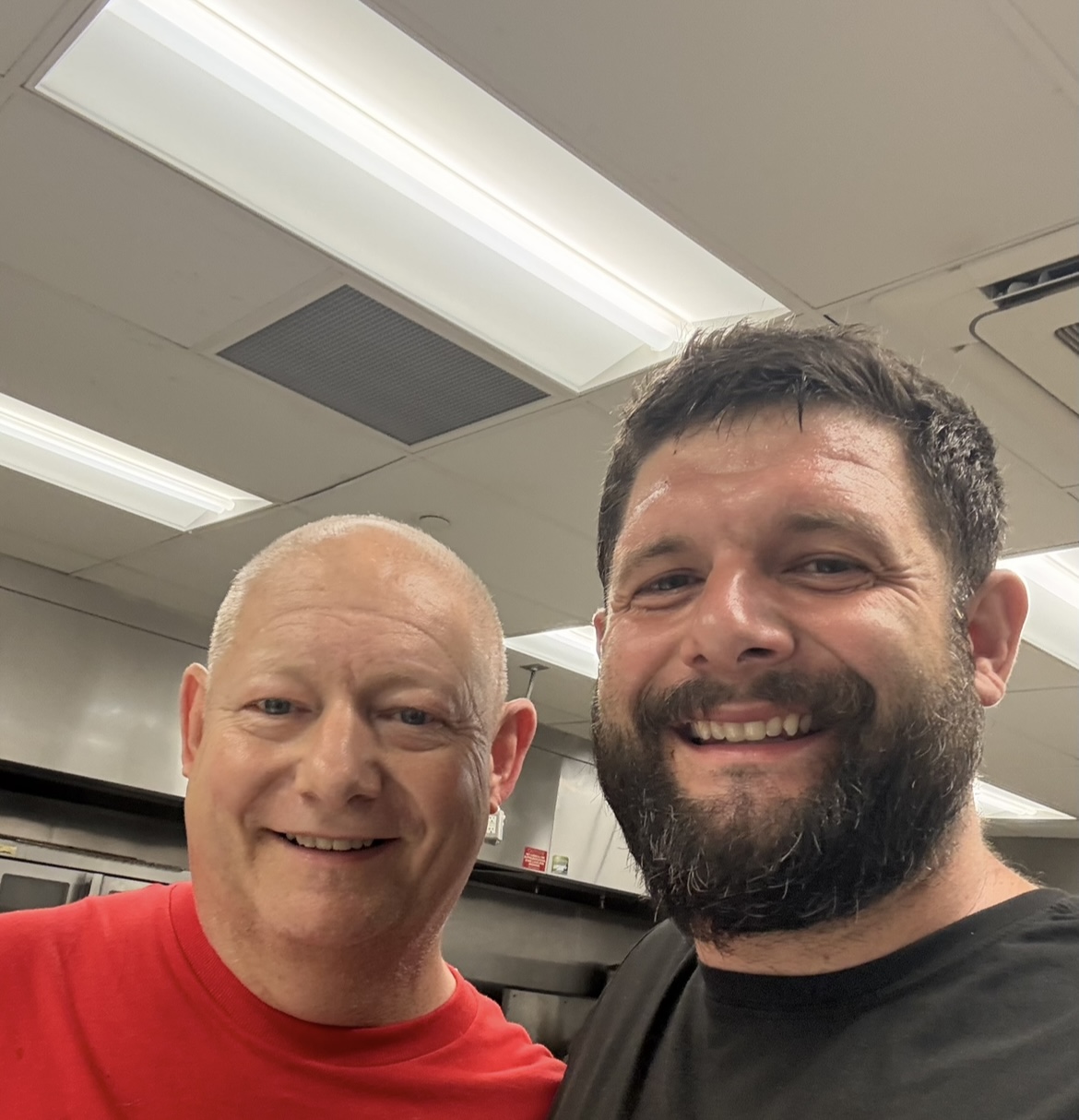 Two smiling men taking a selfie indoors, ceiling lights visible above them.