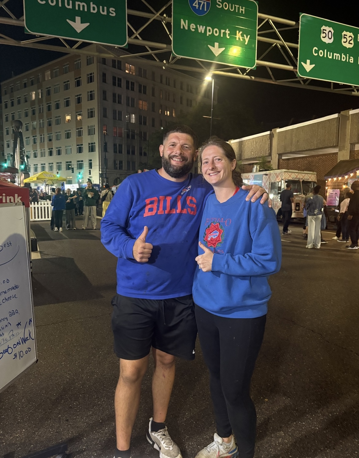 Two people smiling and giving thumbs-up at night in a city street with green highway signs overhead.