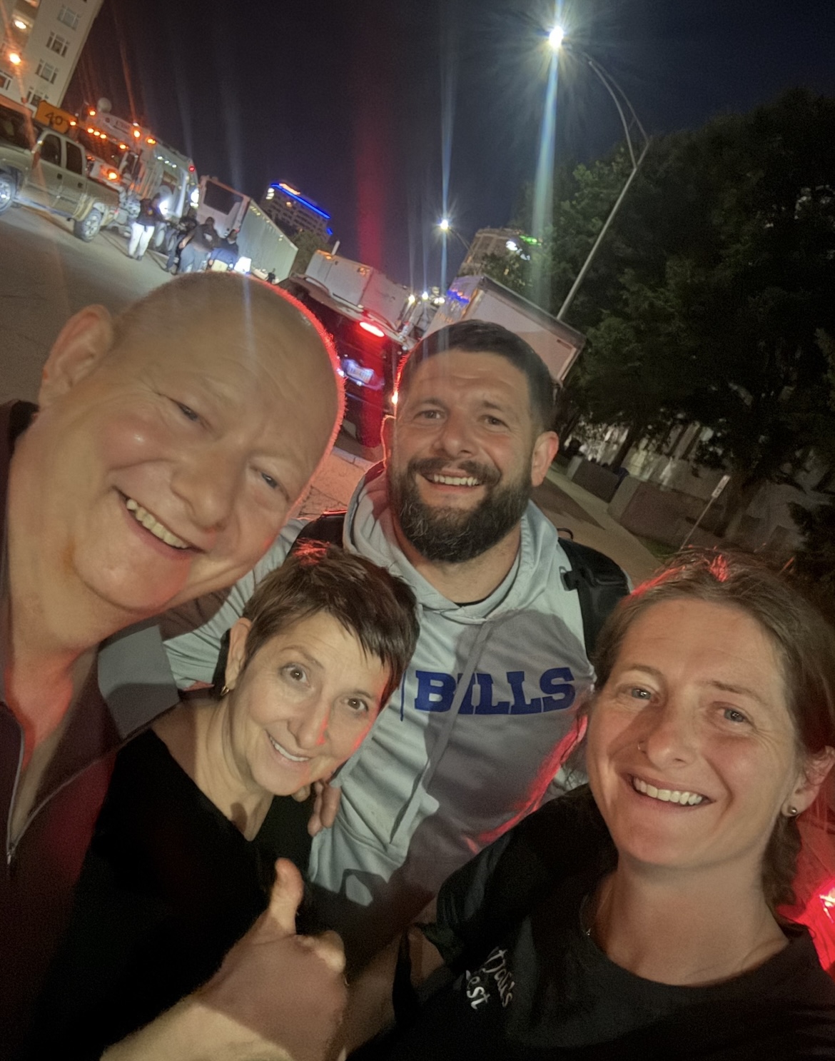 Four people smiling at night on a city street with cars and bright lights in the background.