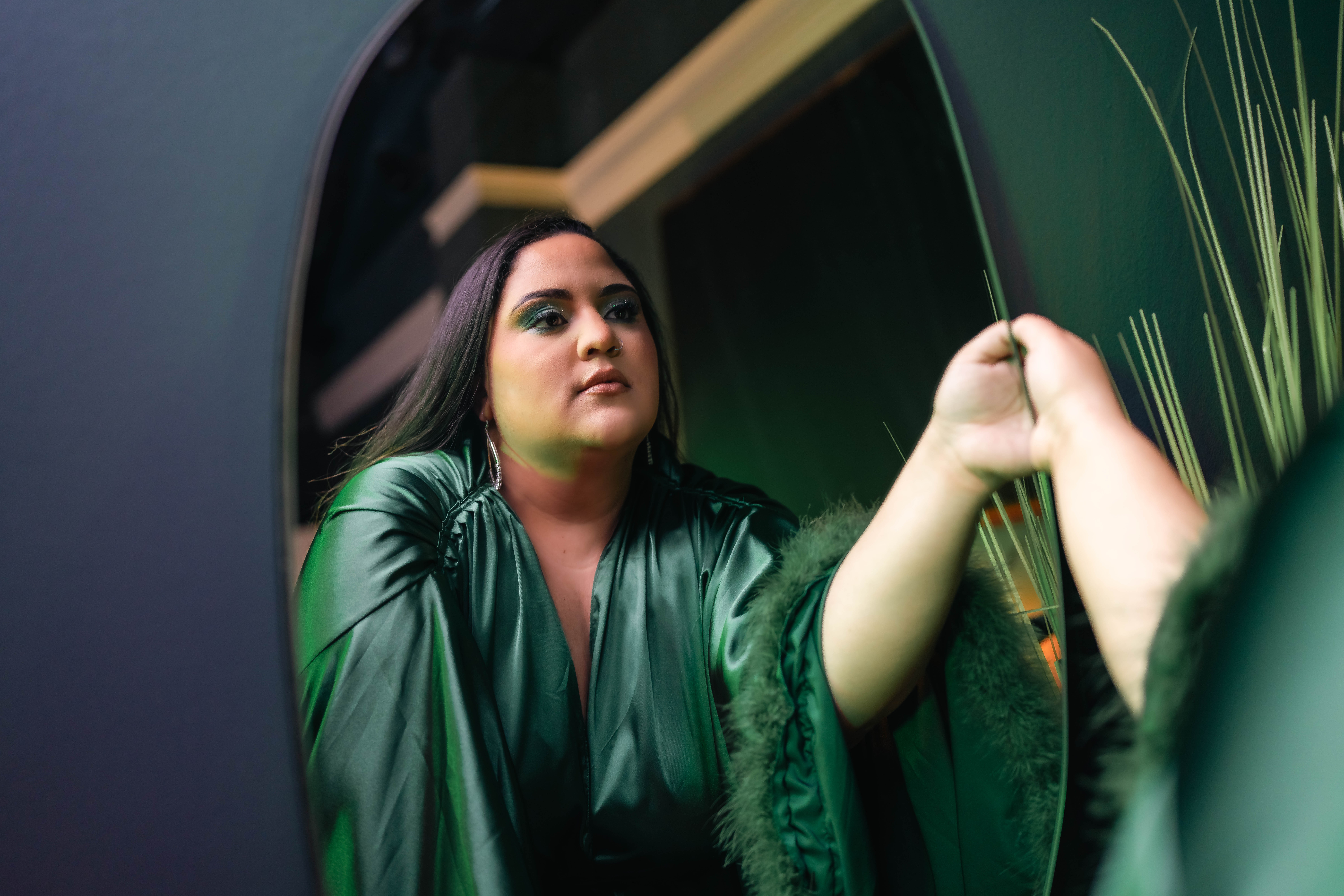 Woman with dark hair and green outfit looking at her reflection in a mirror, holding her hand up, with a green background.