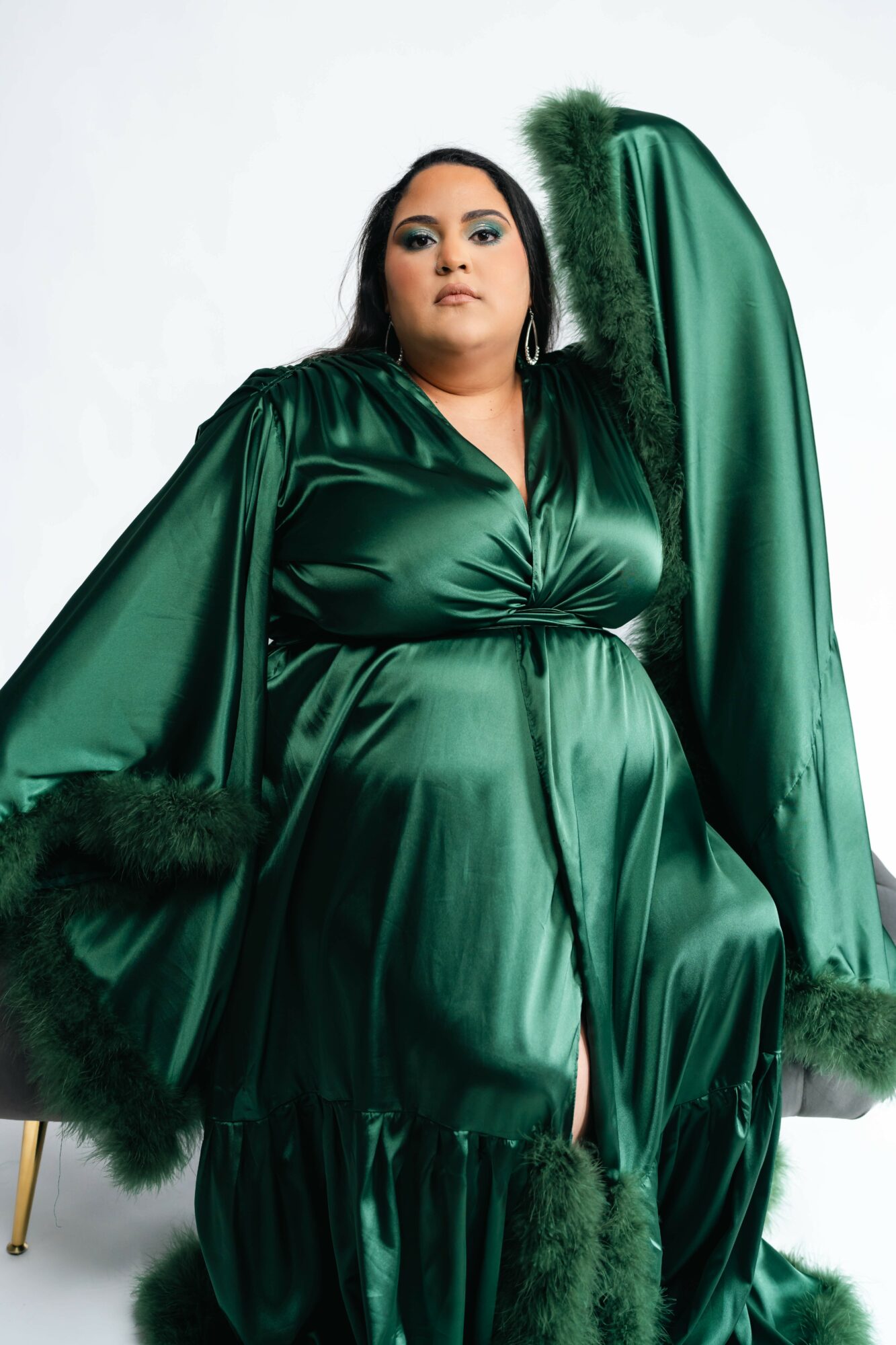 Woman in a shiny green dress with long sleeves and fur trim, standing against a white background.