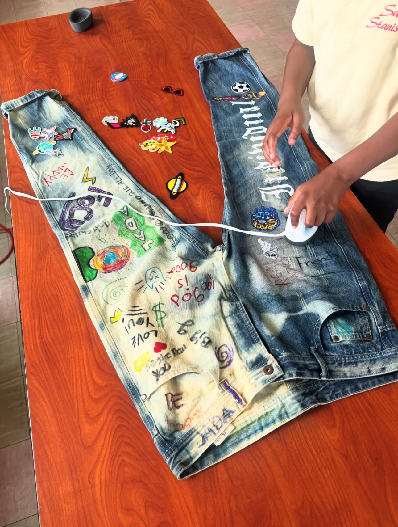 Two pairs of jeans on a table decorated with stickers, drawings, and a person applying a sticker to one jeans leg.